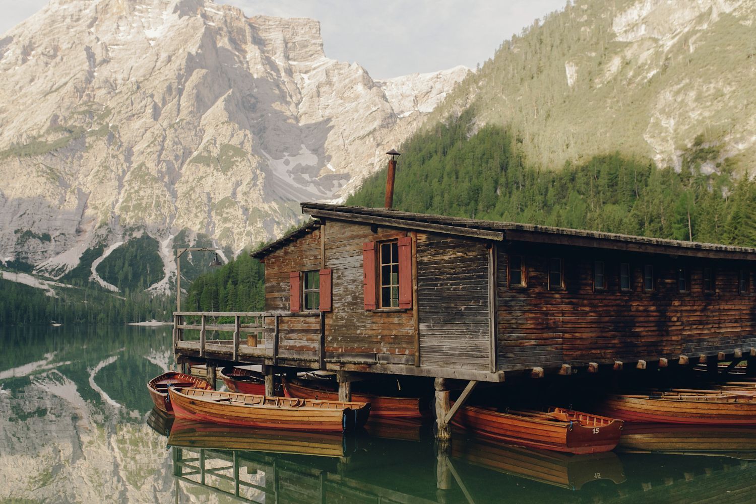 Ein hölzernes Bootshaus an einem See mit Ruderbooten. Im Hintergrund spiegeln sich Berge und Bäume im Wasser.