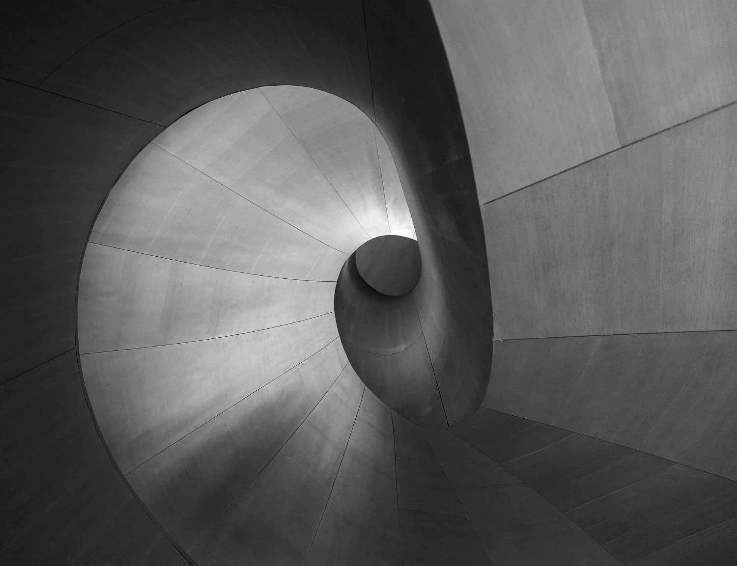 Wendeltreppe aus grauem Beton, die sich nach unten erstreckt und in deren Mitte ein heller Punkt liegt.