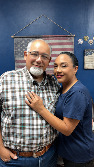 Two people smiling indoors, standing close together in front of an American flag wall hanging.