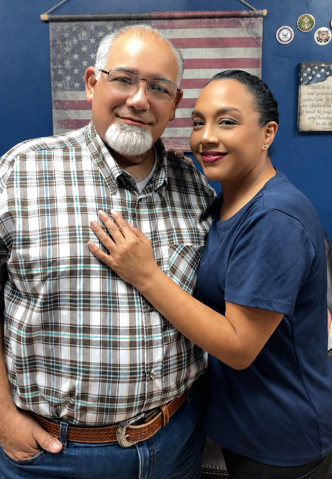 Two people smiling indoors, standing close with one hand on the man's chest.