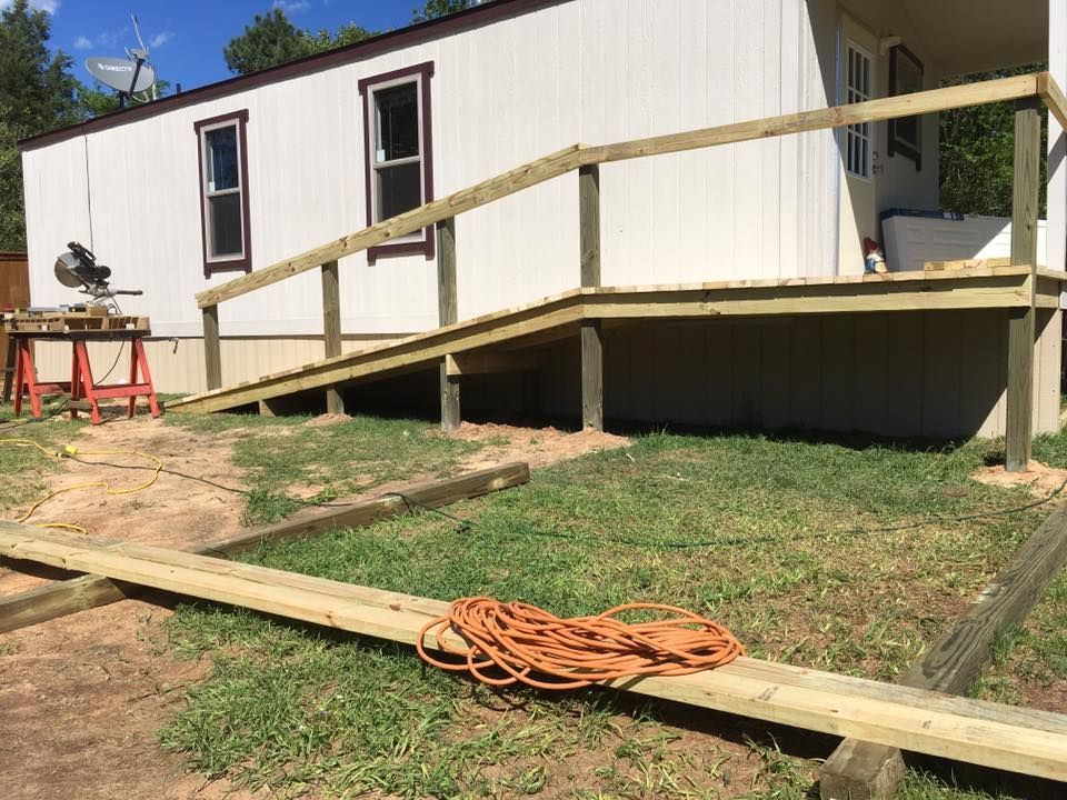 A wooden wheelchair ramp under construction leads to the porch of a white mobile home, with a miter saw set up nearby.