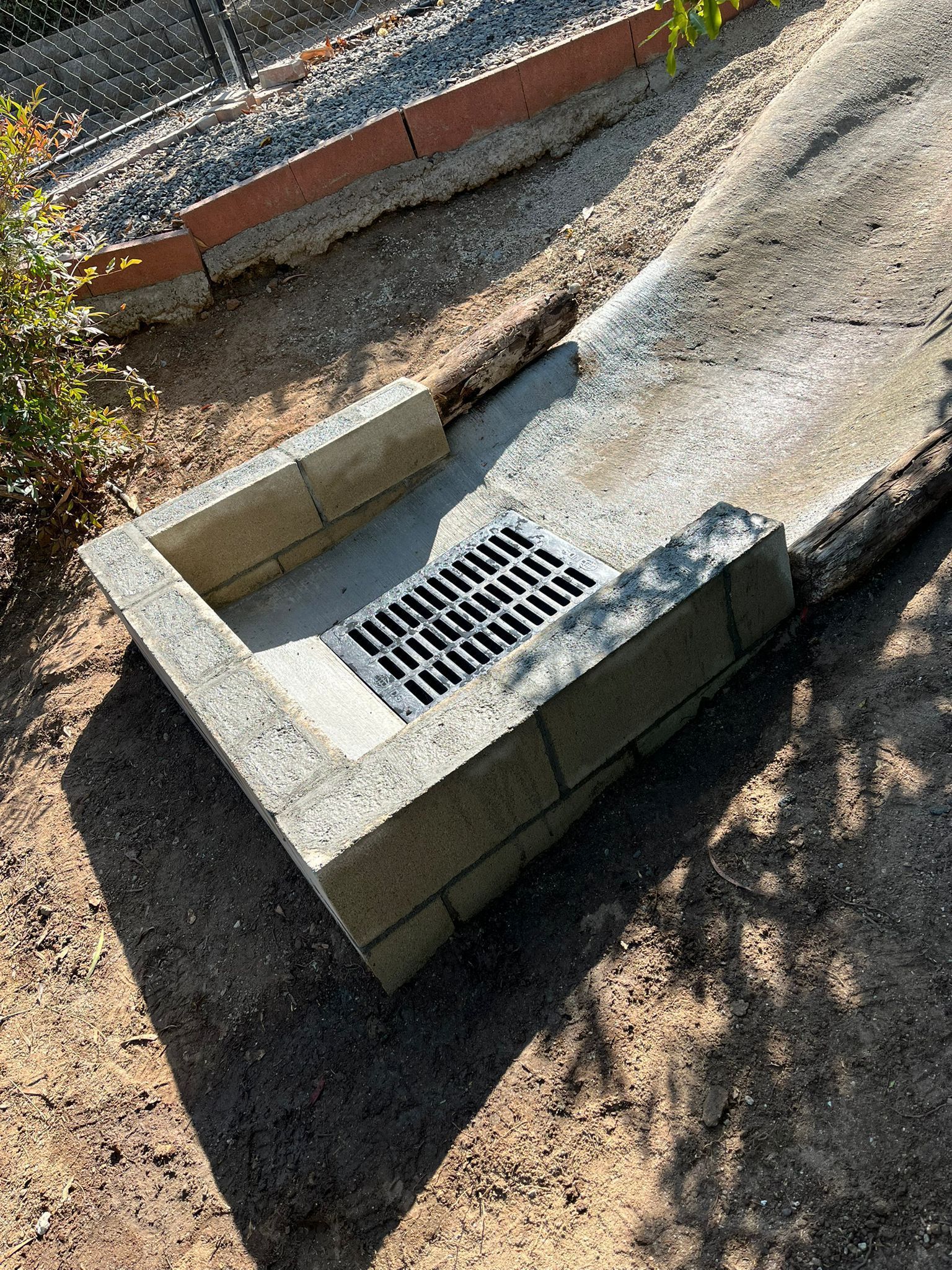 Concrete drainage pit with metal grate beside a sloped gravel path and brick edging
