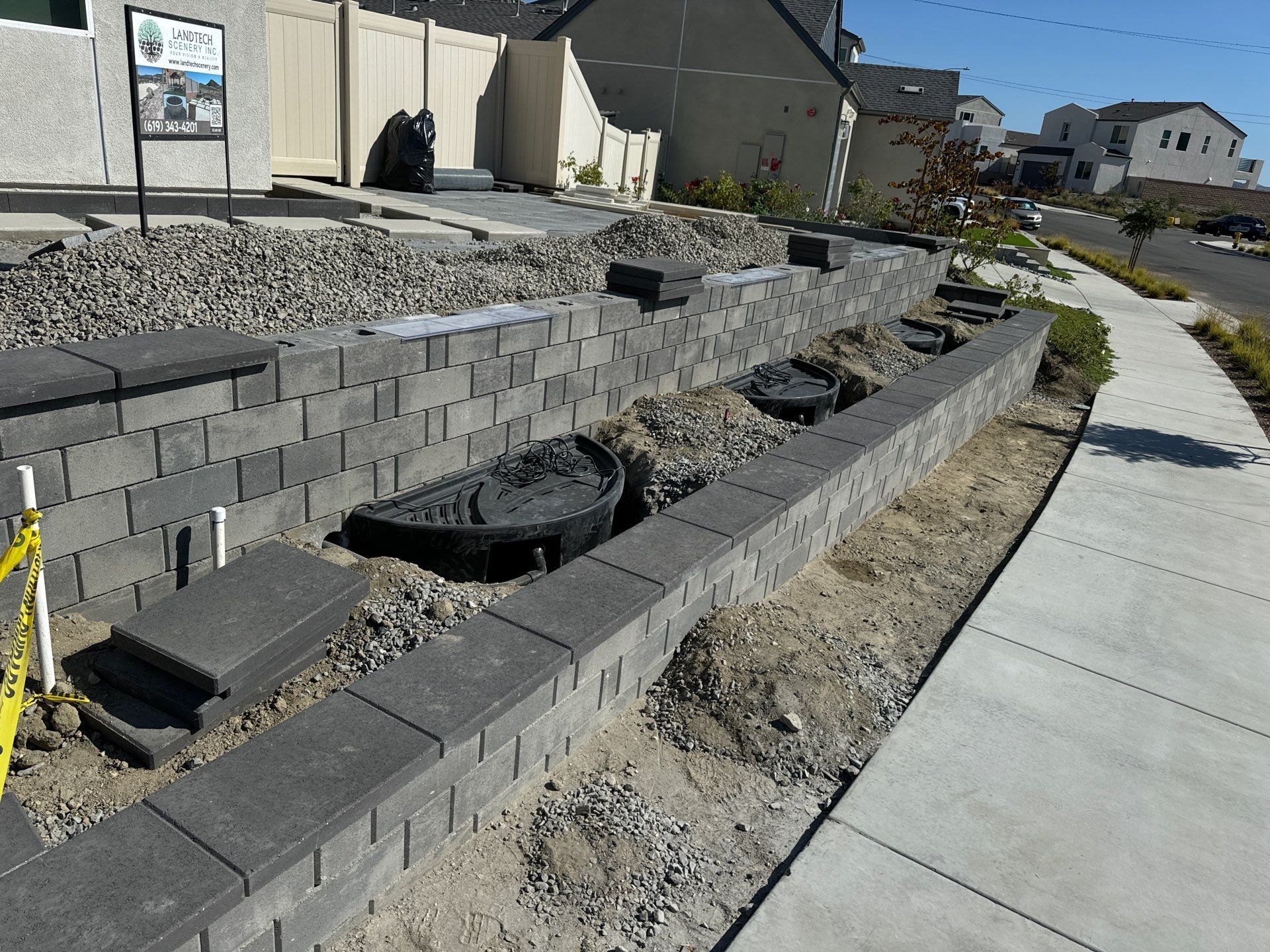 Retaining wall under construction with gray blocks, gravel, and concrete near a sidewalk.