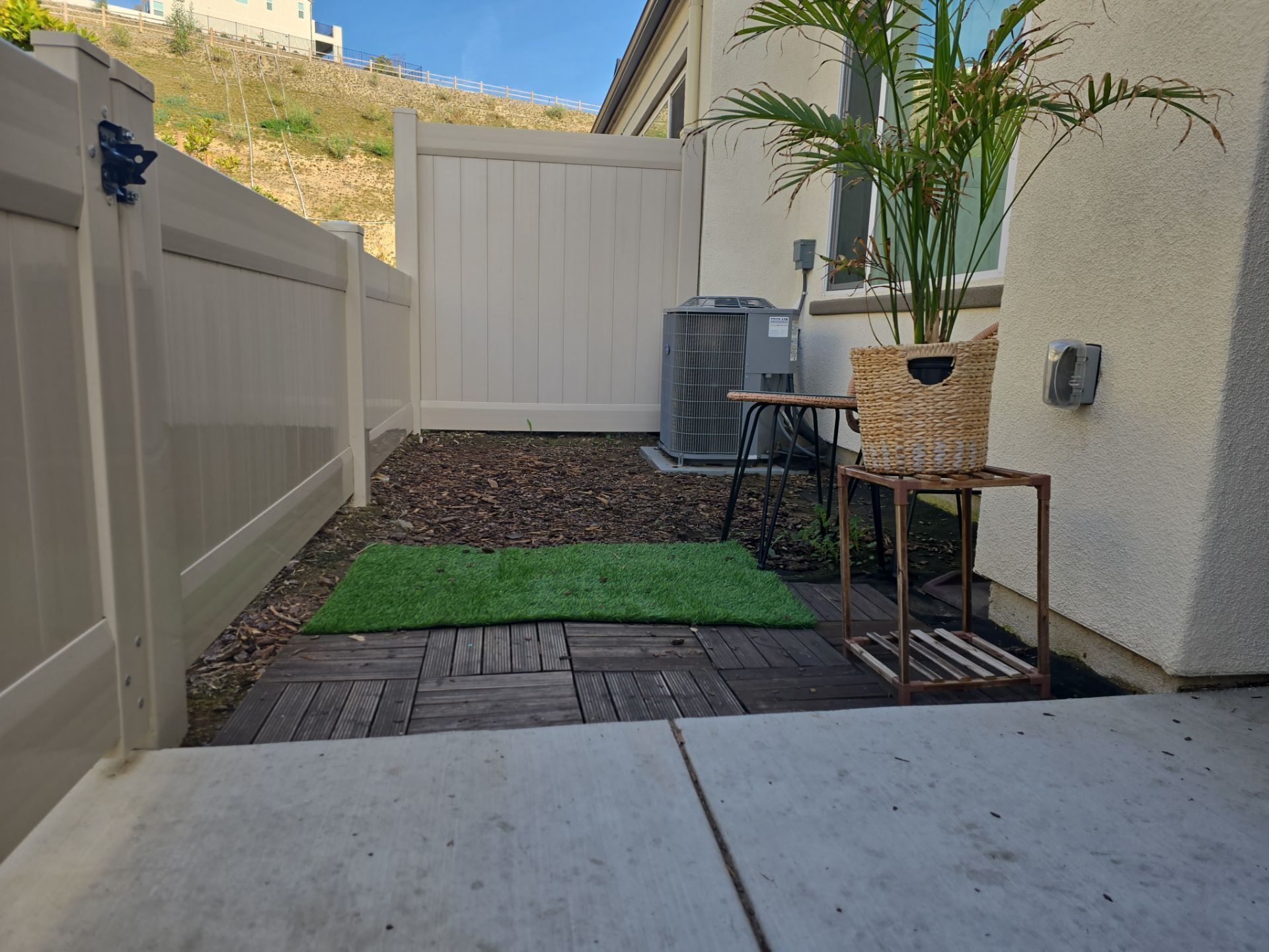 Fenced patio area with artificial grass, a potted plant, small table, and an AC unit.