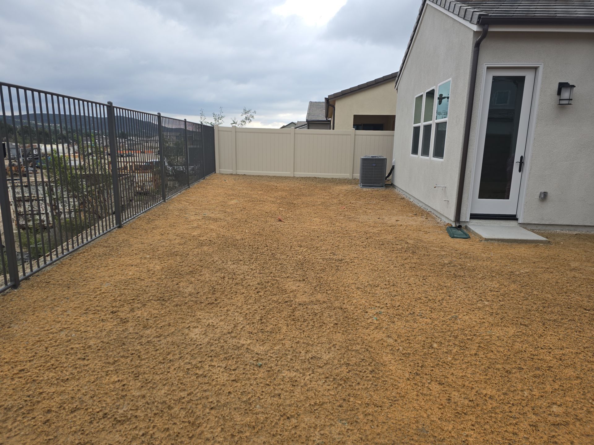 Gravel-covered backyard with a black fence, beige fence, and a house with a door and windows. Overcast sky.