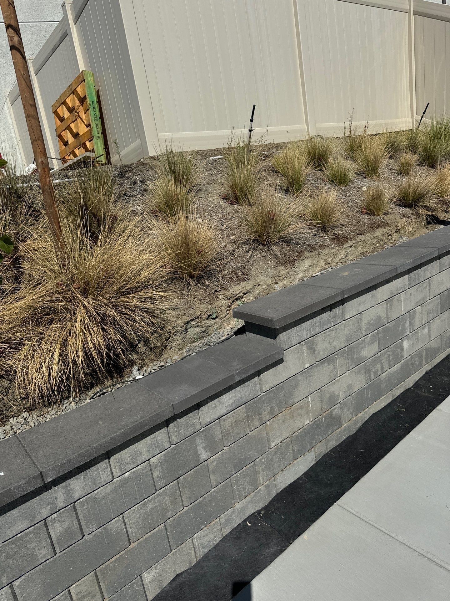 A gray brick retaining wall with a dark cap, with dry grass and a white fence in the background.