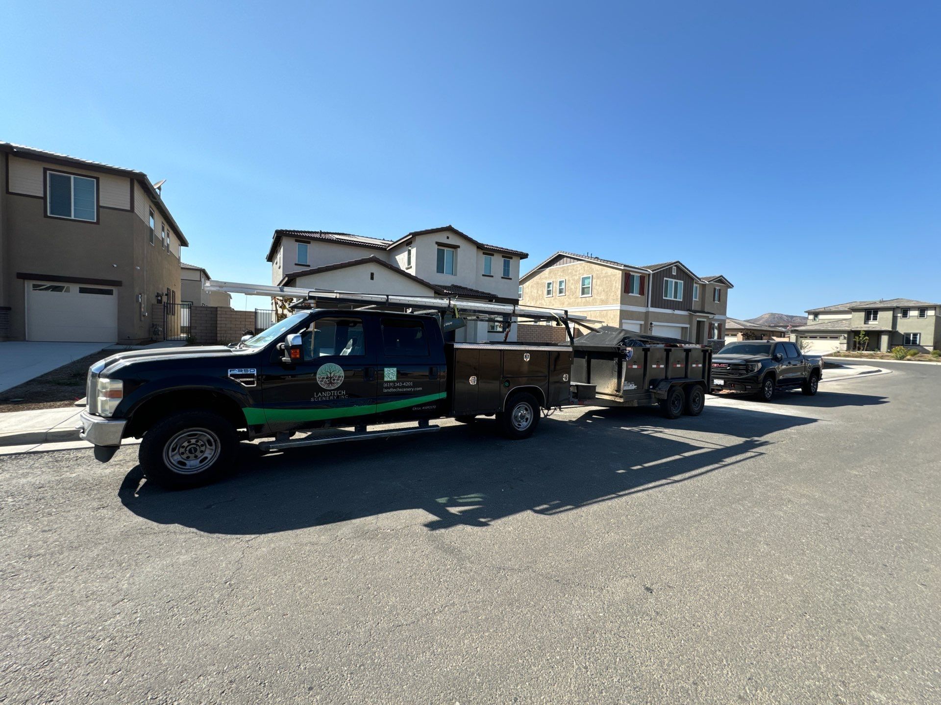 Dark truck with trailer parked in a suburban street, houses in background under a blue sky.