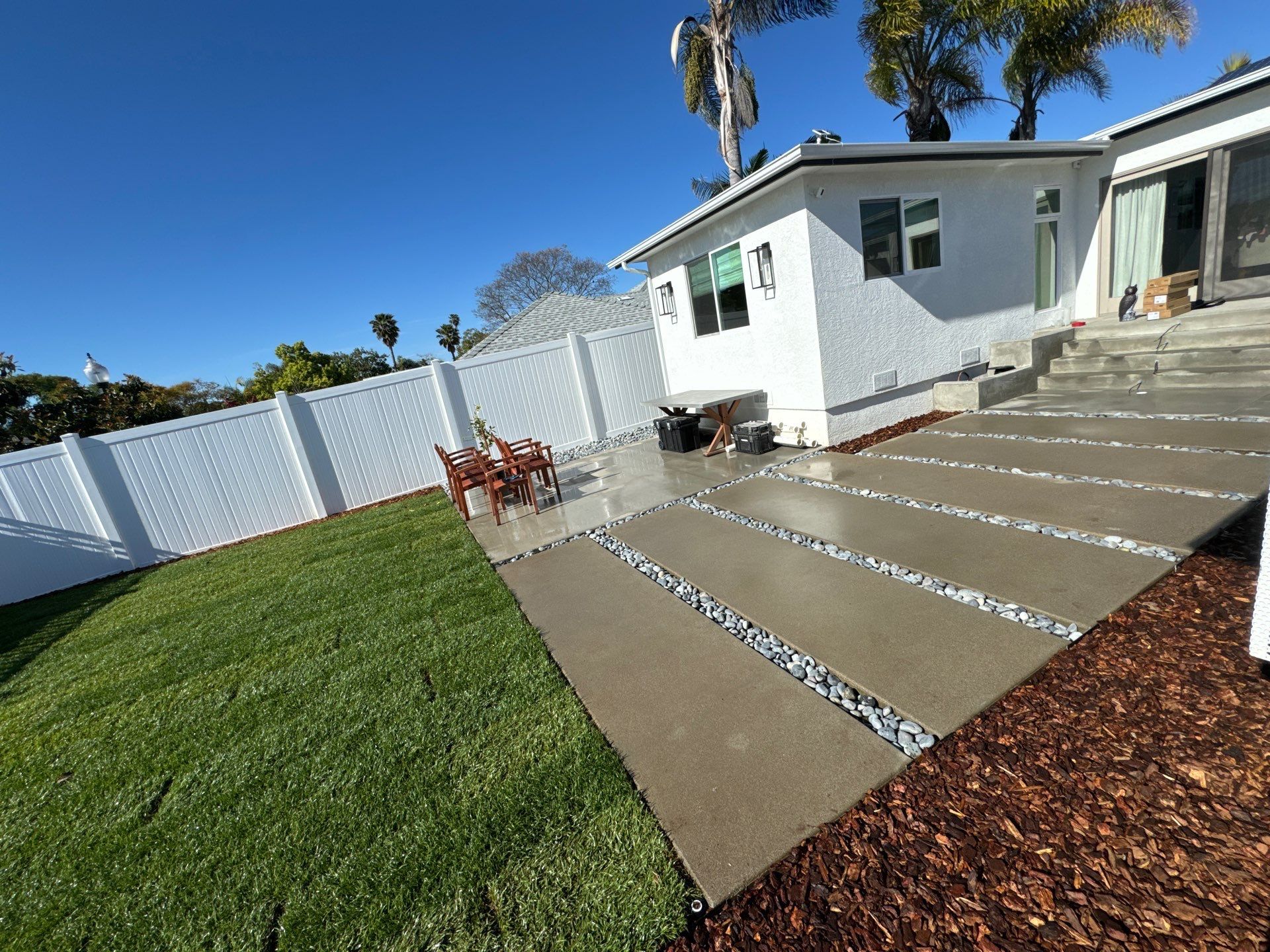 Backyard with white fence, green grass, concrete walkway with pebbles, and white house.