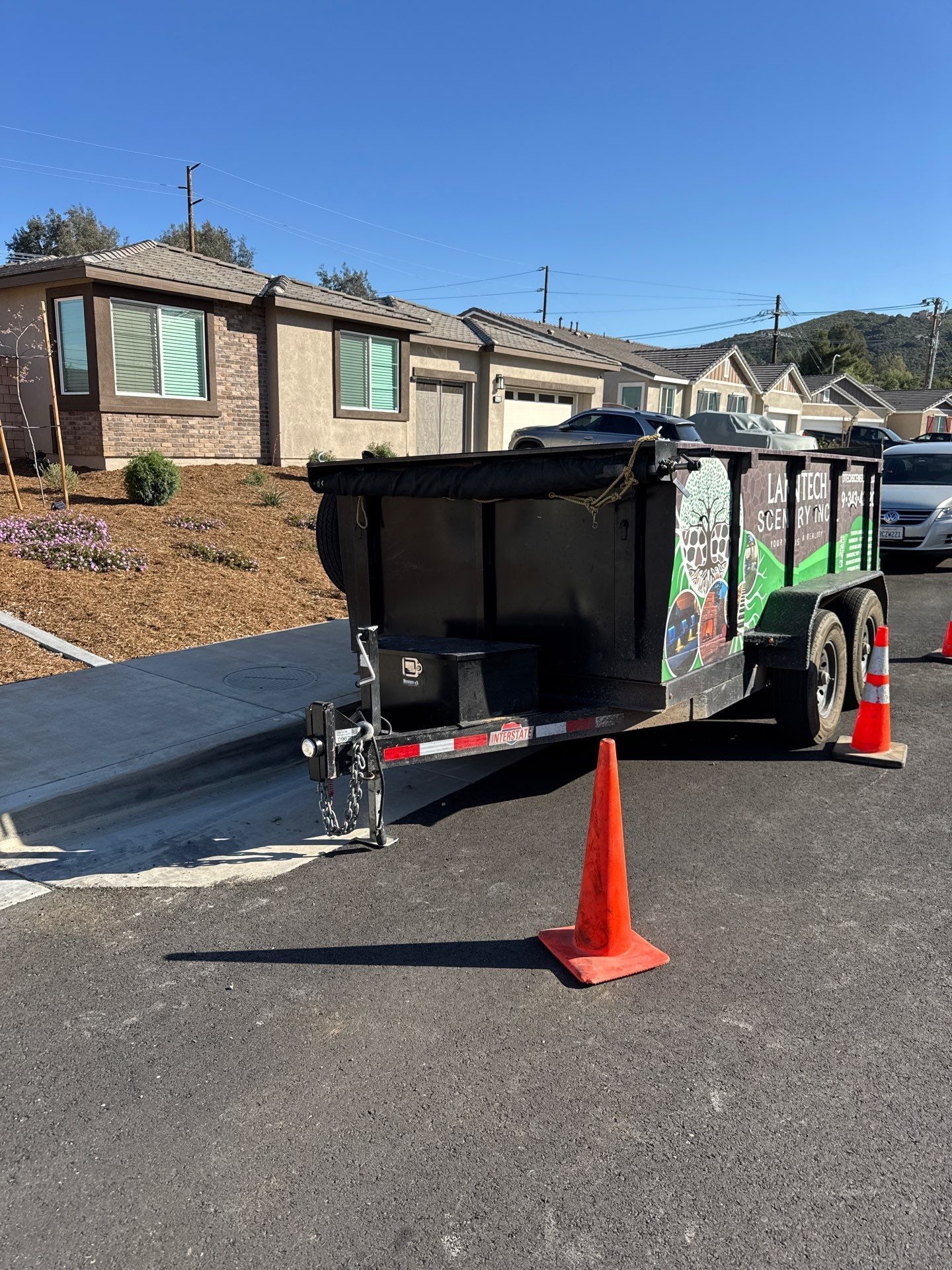 A dumpster trailer on a paved road, orange traffic cone in front. Houses and blue sky in the background.
