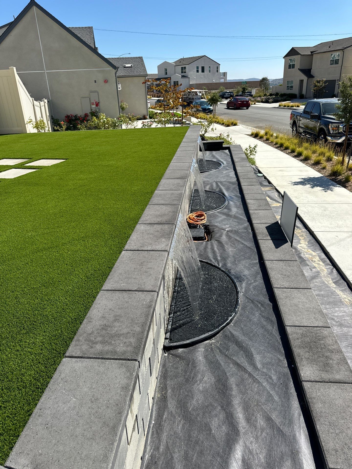 Long rectangular planter box with gray bricks, filled with black fabric, next to artificial green grass and sidewalk.