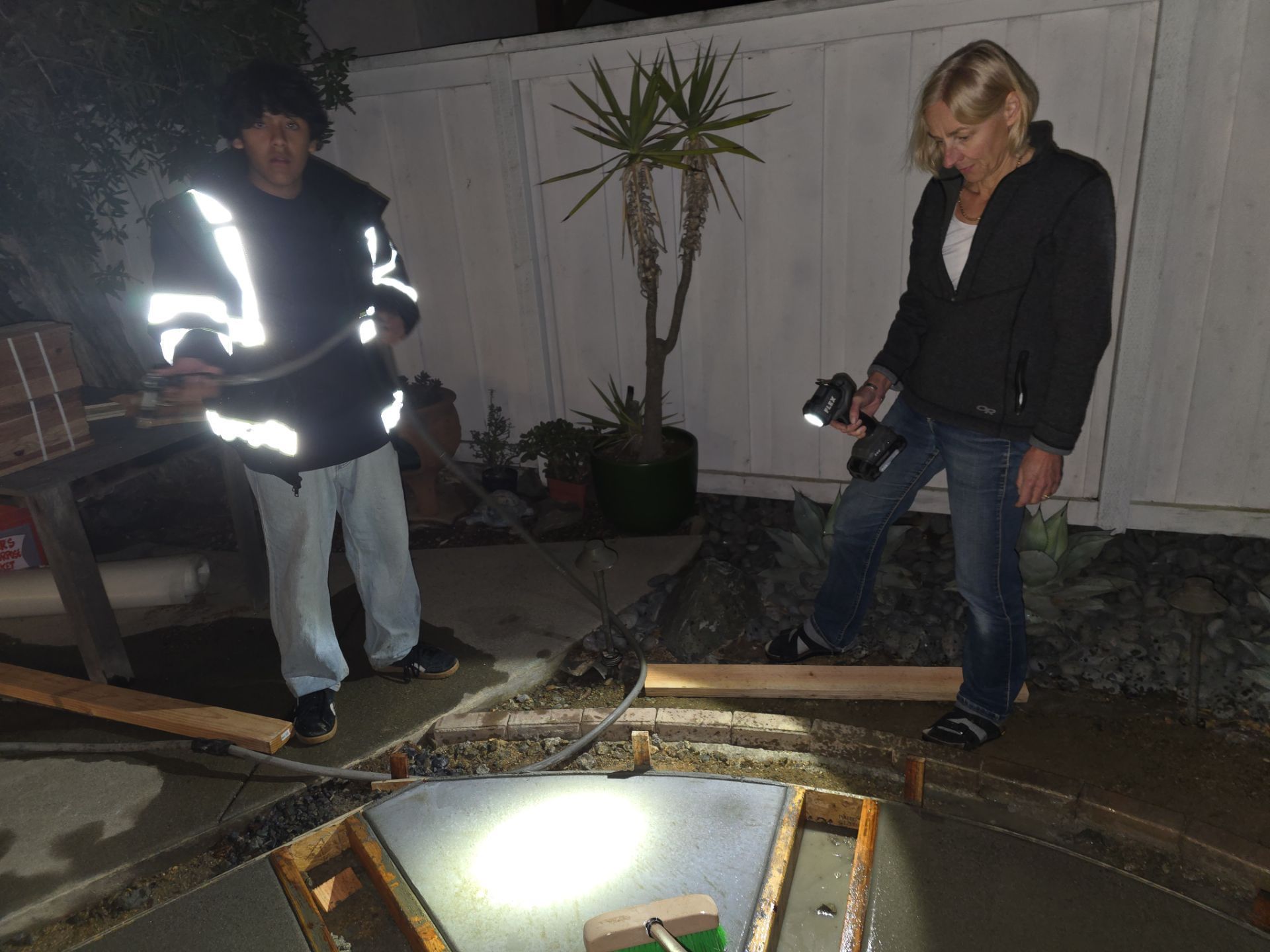 Two people inspecting a newly poured concrete project at night, one holding a flashlight and the other a hose.