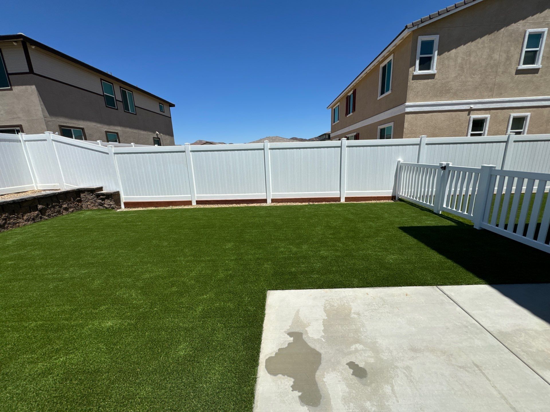 Backyard with artificial green grass, white fence, and beige houses under a blue sky.
