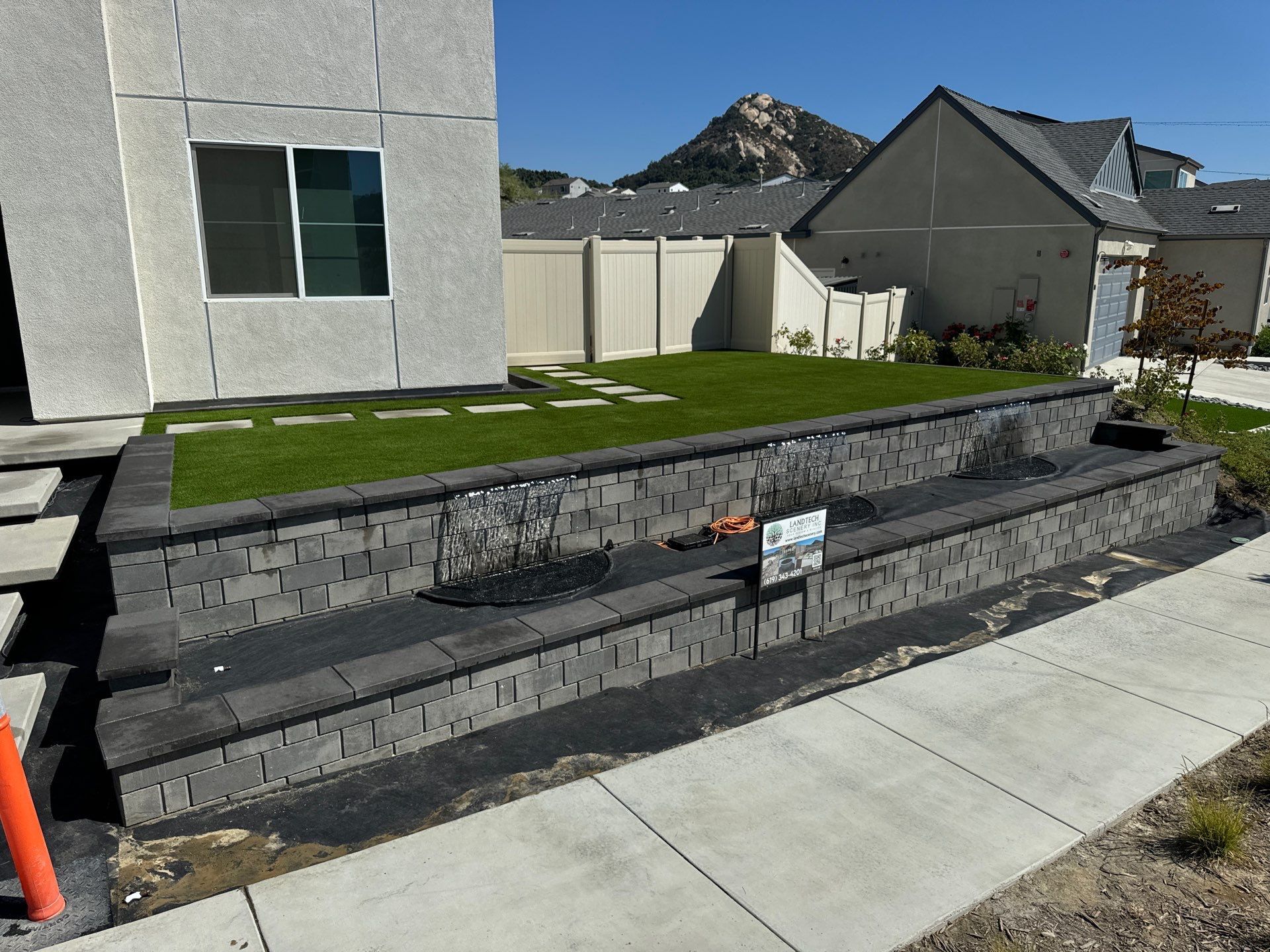 Two-tiered retaining wall with dark pavers, holding a patch of grass. A building and neighborhood are in the background.