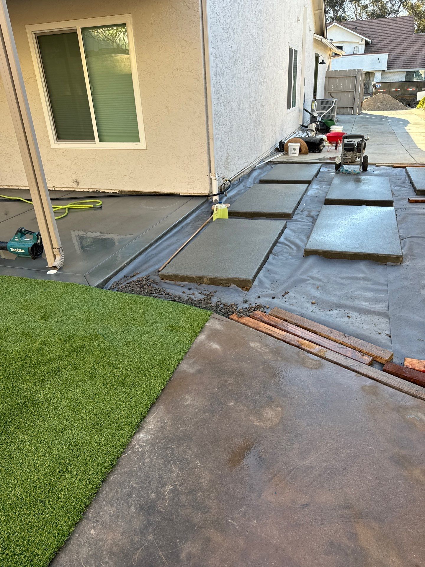 Concrete stepping stones laid on a black fabric in a backyard, next to an artificial turf patch and a house.