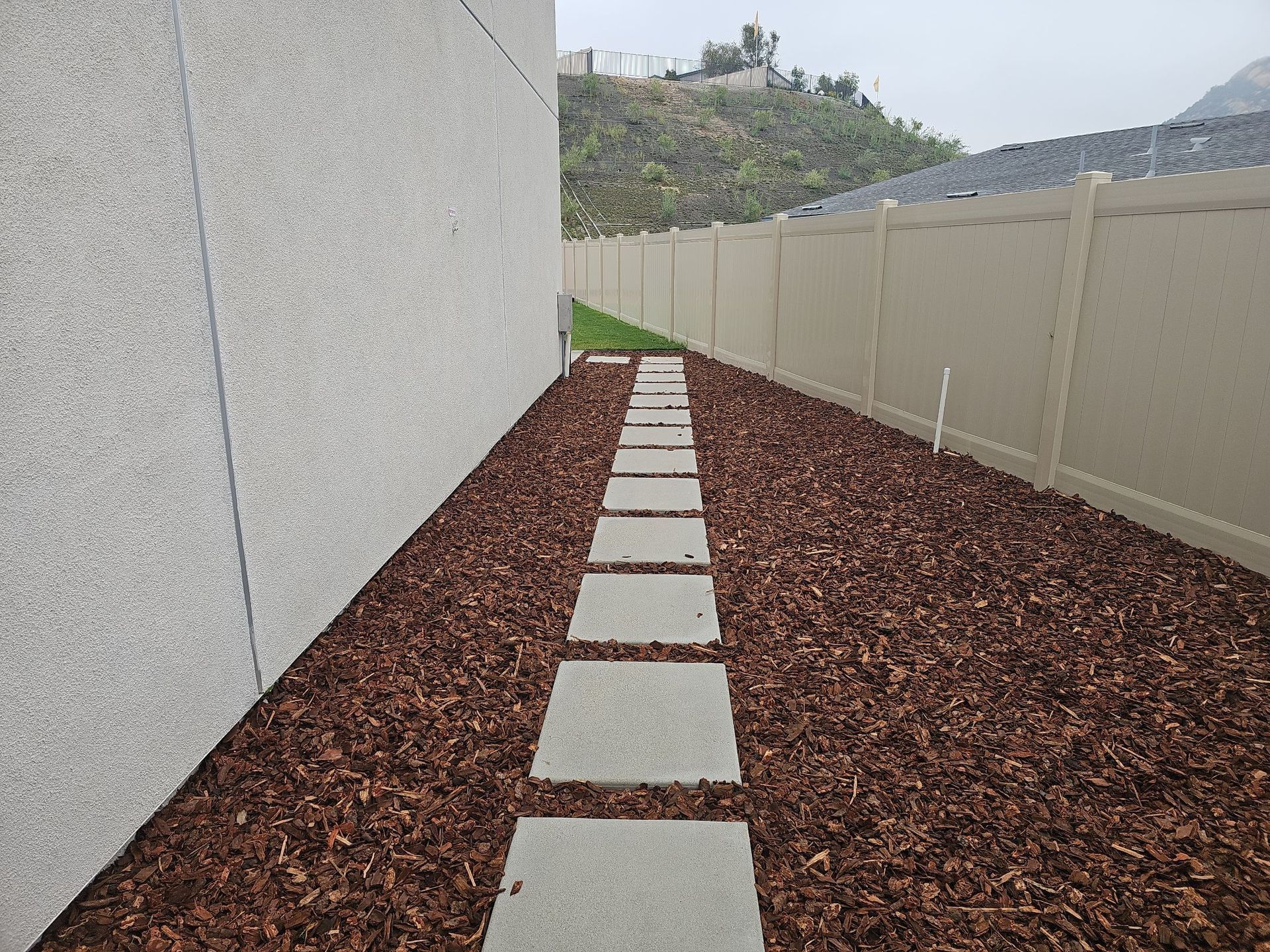 Stone path through wood chips along a building wall and fence, with a hillside in the background.
