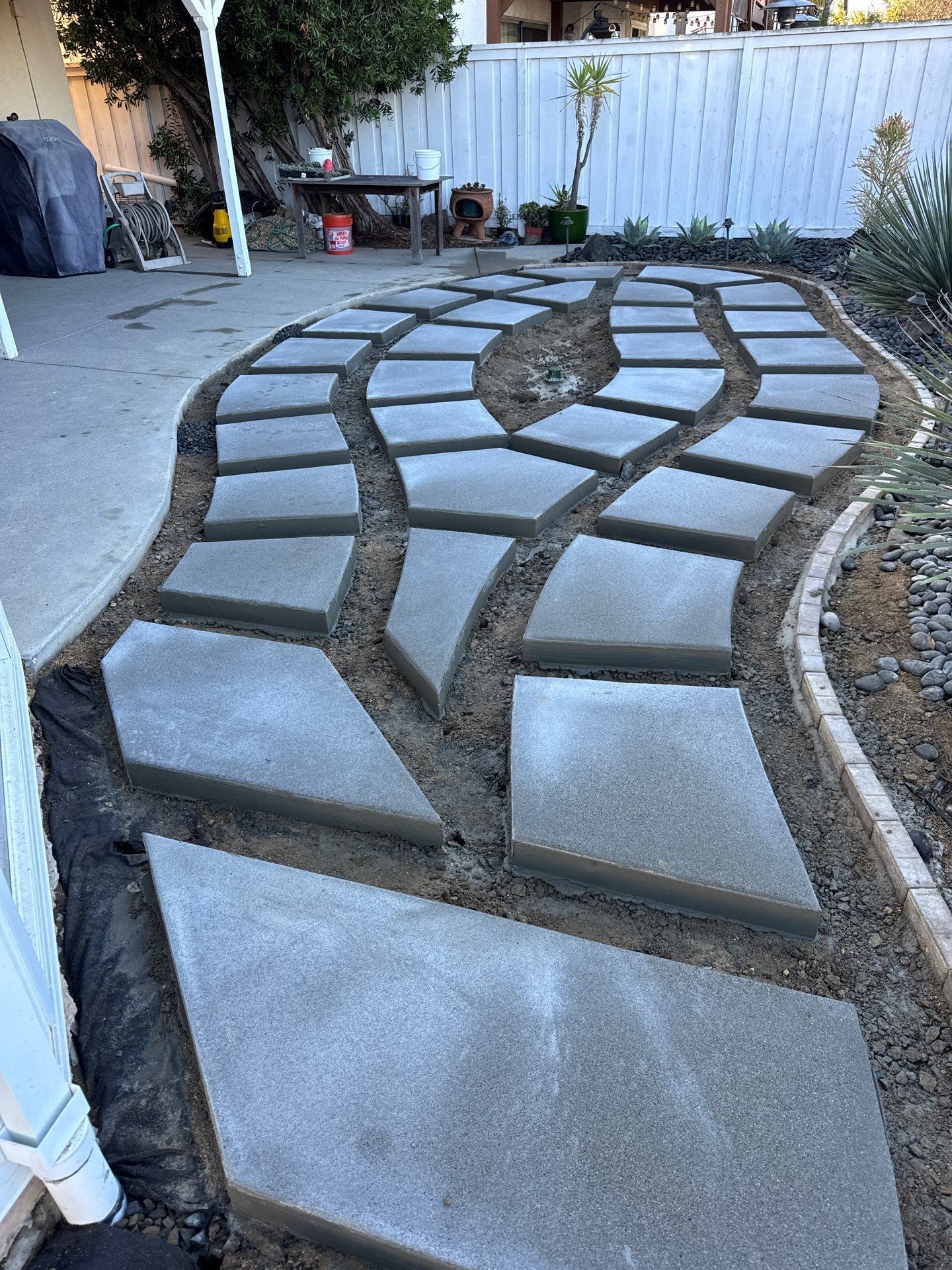 Concrete pavers forming a pathway in a backyard, surrounded by dirt and border, leading toward a white fence.
