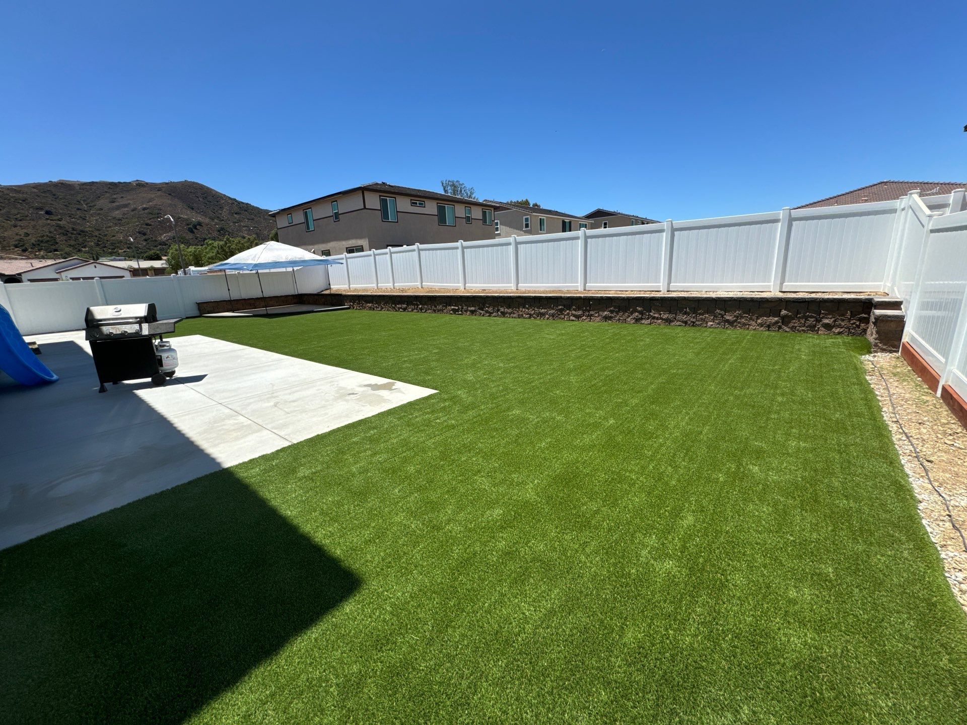 Backyard with green turf, concrete patio, white fence, and a barbecue grill on a sunny day.