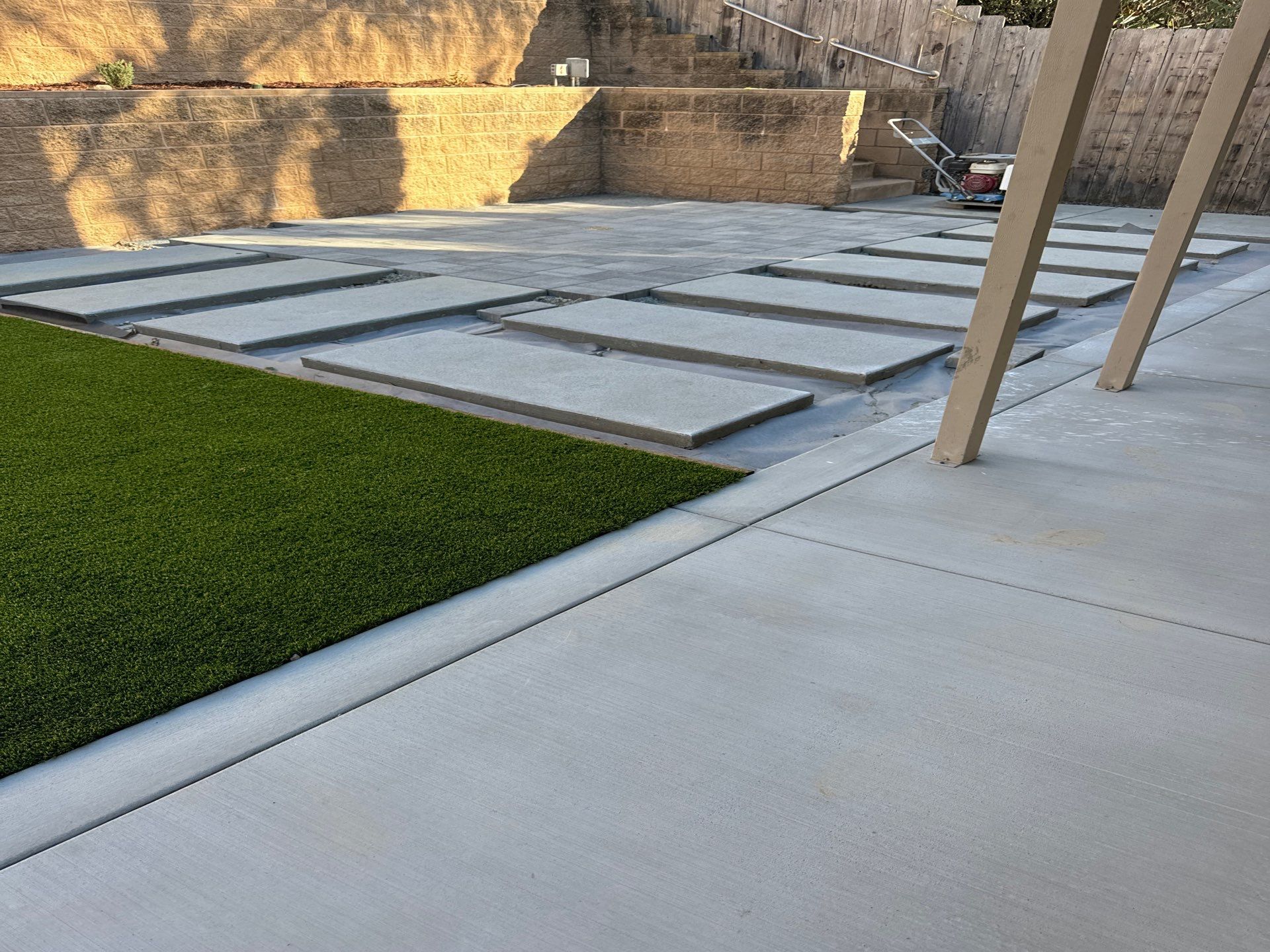 Concrete patio with stepping stones, artificial grass, and retaining wall in a backyard.