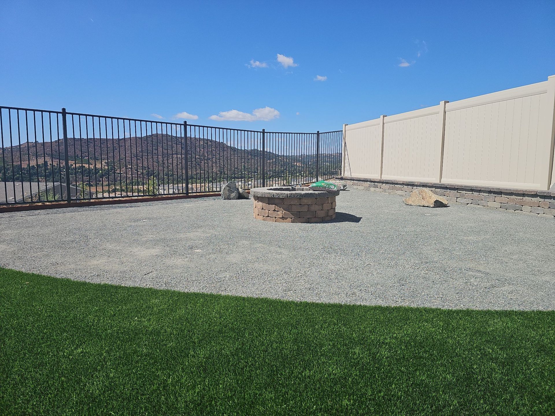 A backyard with a fire pit surrounded by gravel, green lawn, and a fence under a blue sky.