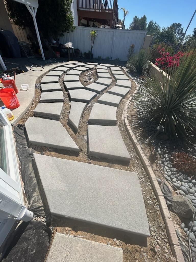 Concrete pavers form a curved path in a backyard with landscaping and a white fence.