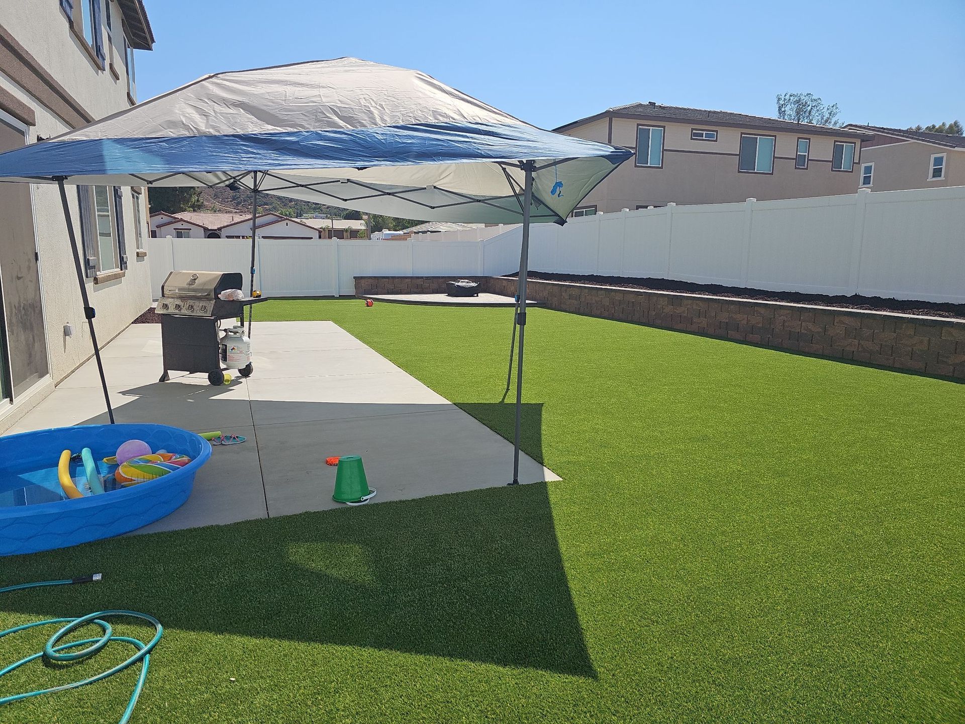 Backyard with canopy, grill, and kiddie pool on artificial grass, sunny day.
