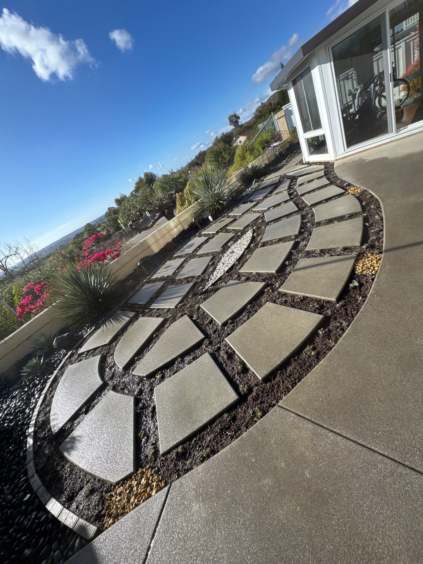 A curved stone walkway bordered by dark gravel and plants, leading to a house with a view.
