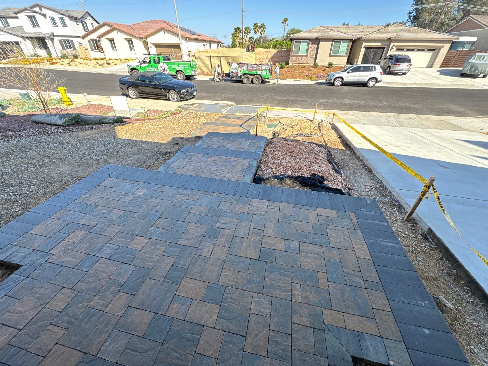Brick pavers being installed on a driveway, with construction vehicles and houses in the background.
