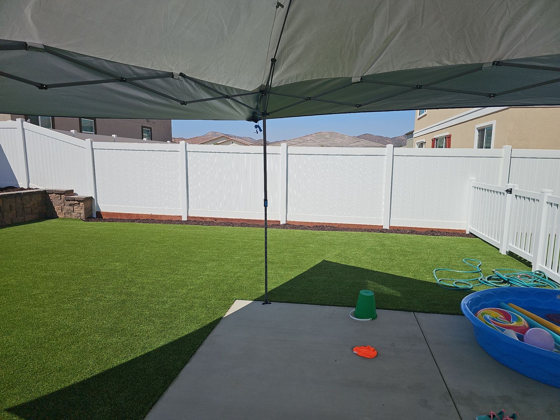 Backyard with artificial grass, white fence, patio, and a canopy. Toys scattered; mountains in the distance.