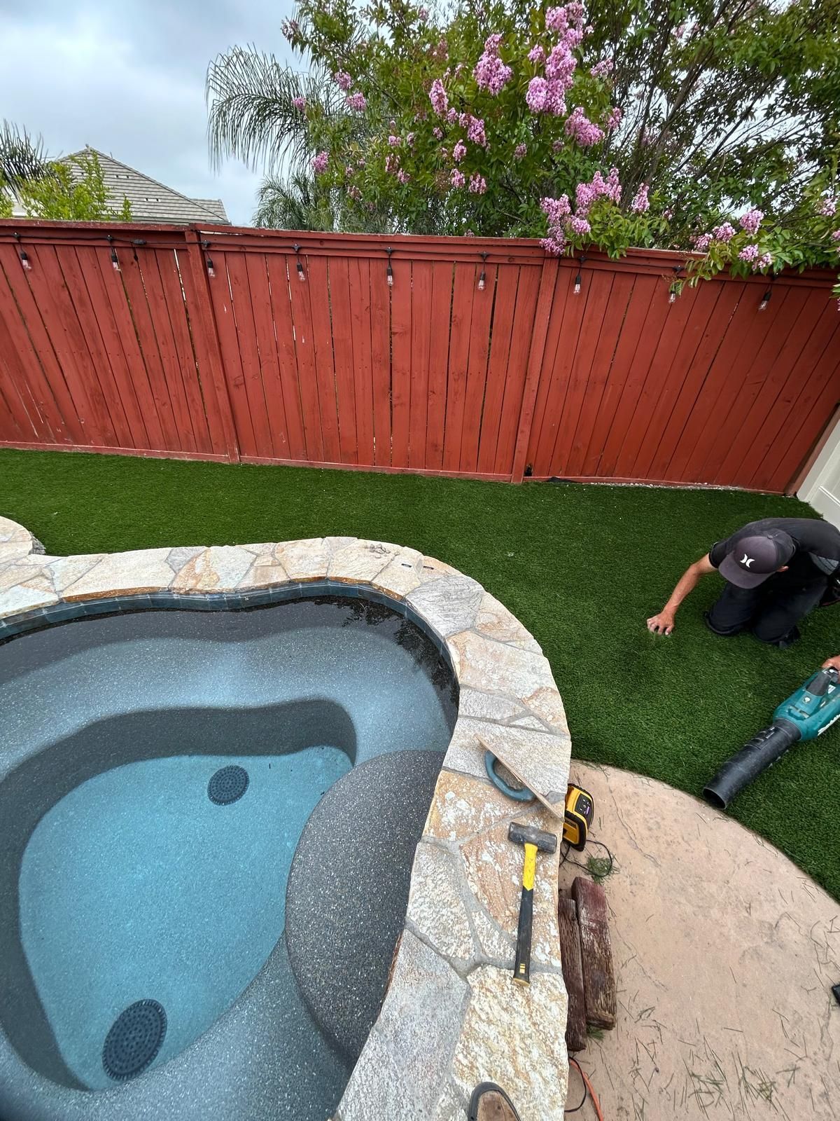 Backyard scene: jacuzzi, red fence, artificial turf, and a person working with a leaf blower.