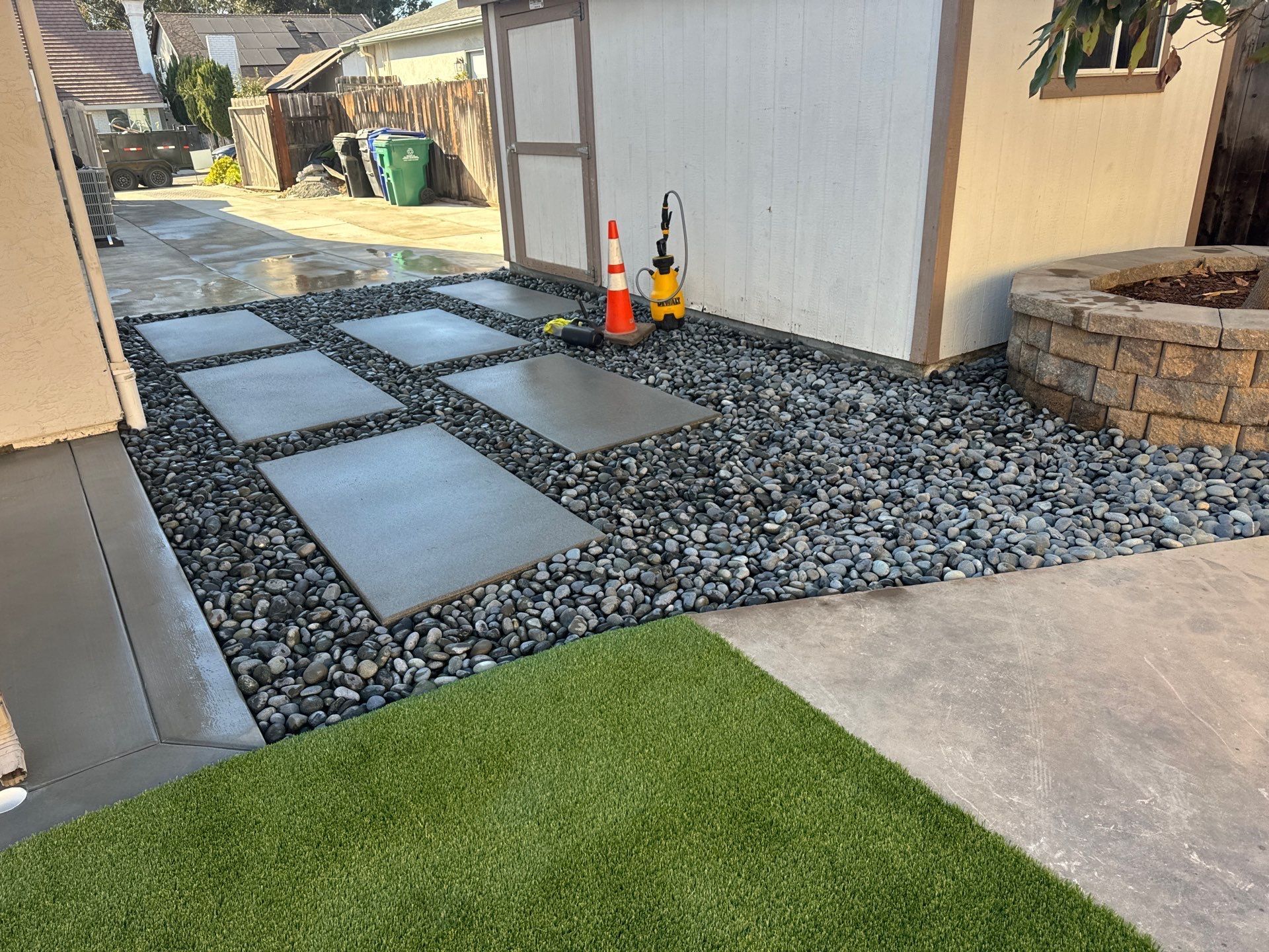 Concrete pavers on a rock bed, next to artificial turf and a shed. A fire pit is on the right.