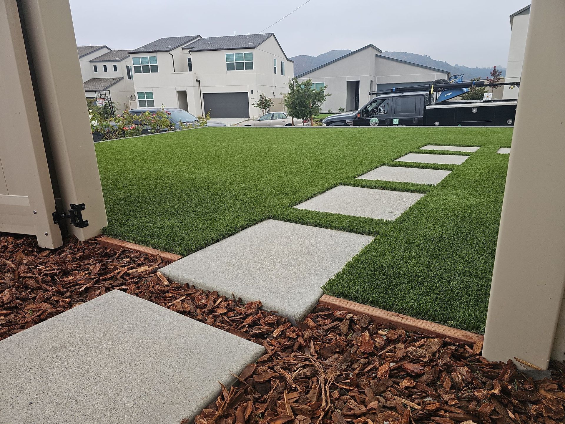 Stone path through artificial lawn, view from gate. Houses and mountains in the background.