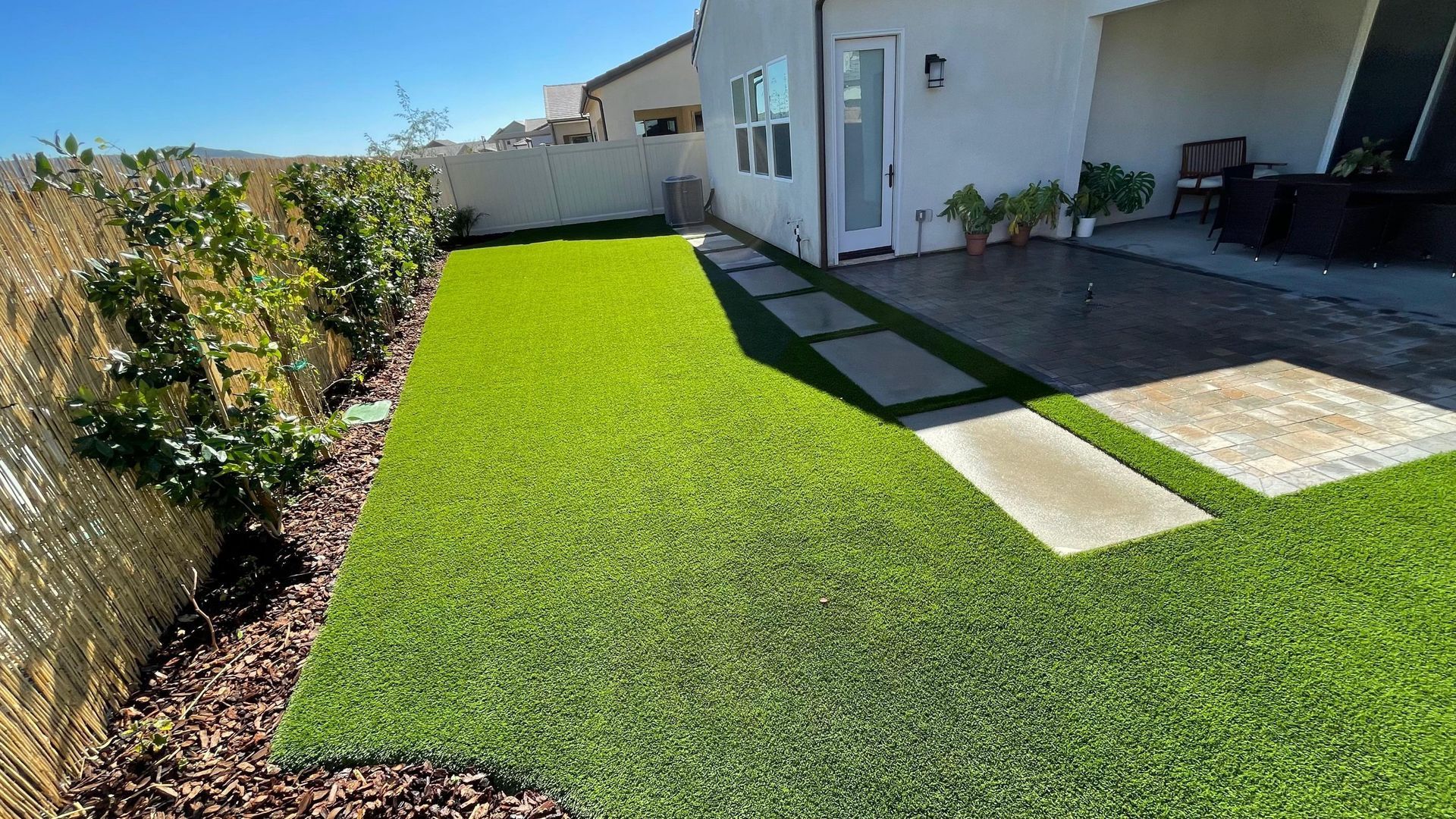 Backyard with artificial green turf, stone path, white house, and bamboo fence.
