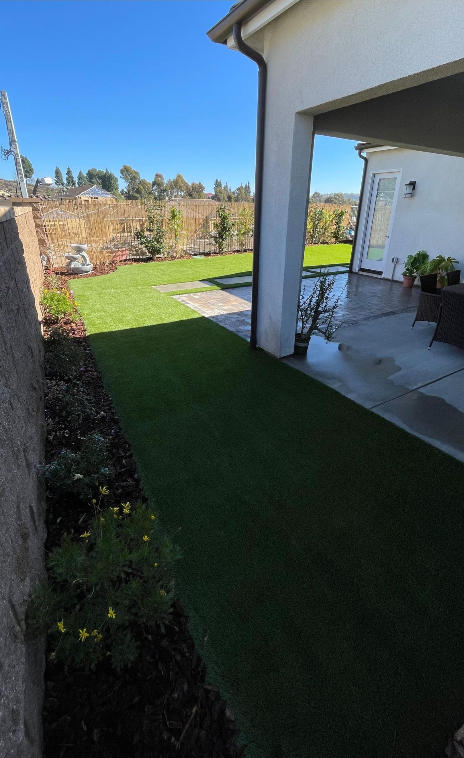A backyard with green turf and a small garden next to a beige house, under a clear blue sky.