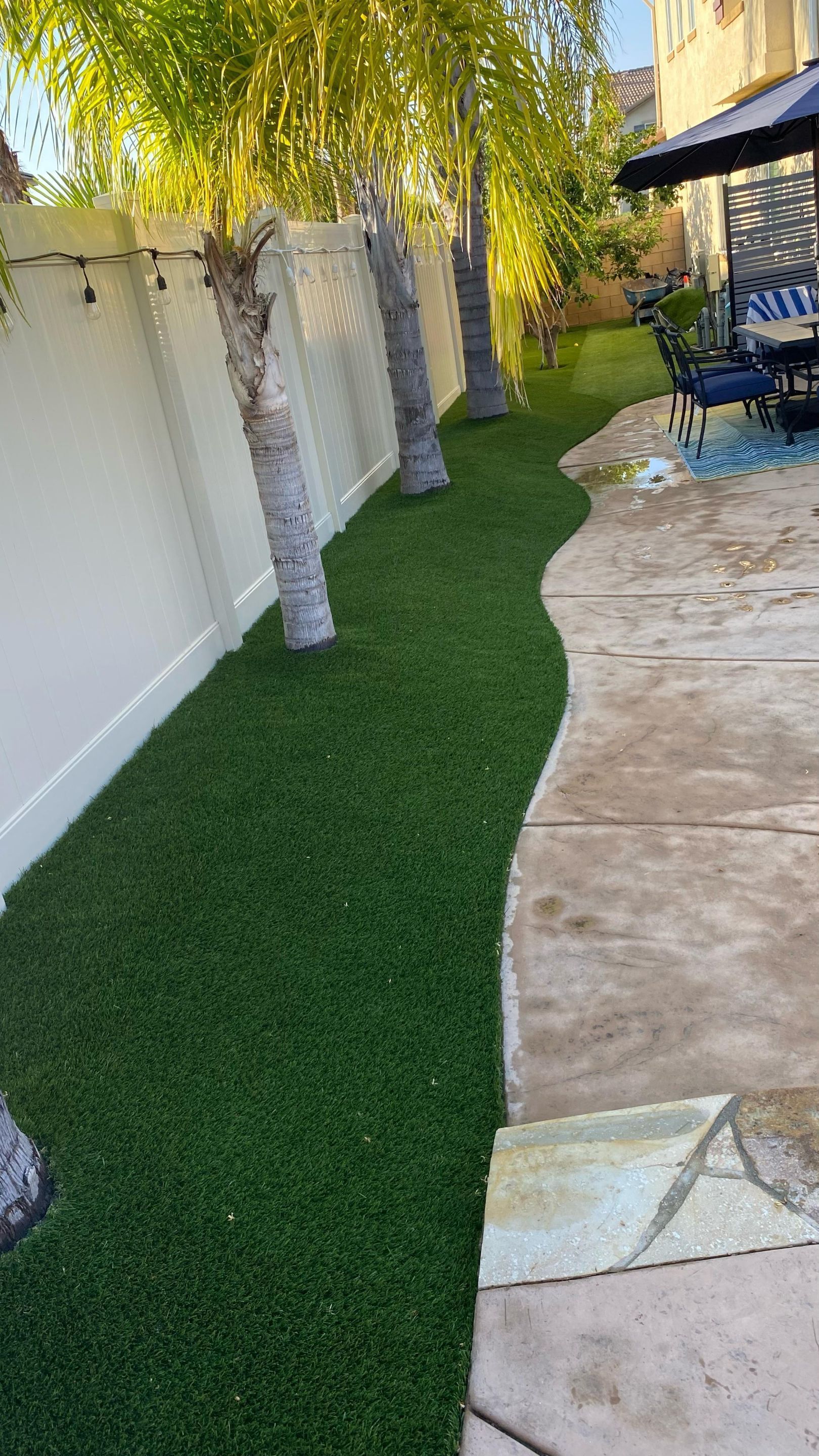 Green artificial turf alongside a white fence and concrete patio, with palm trees.