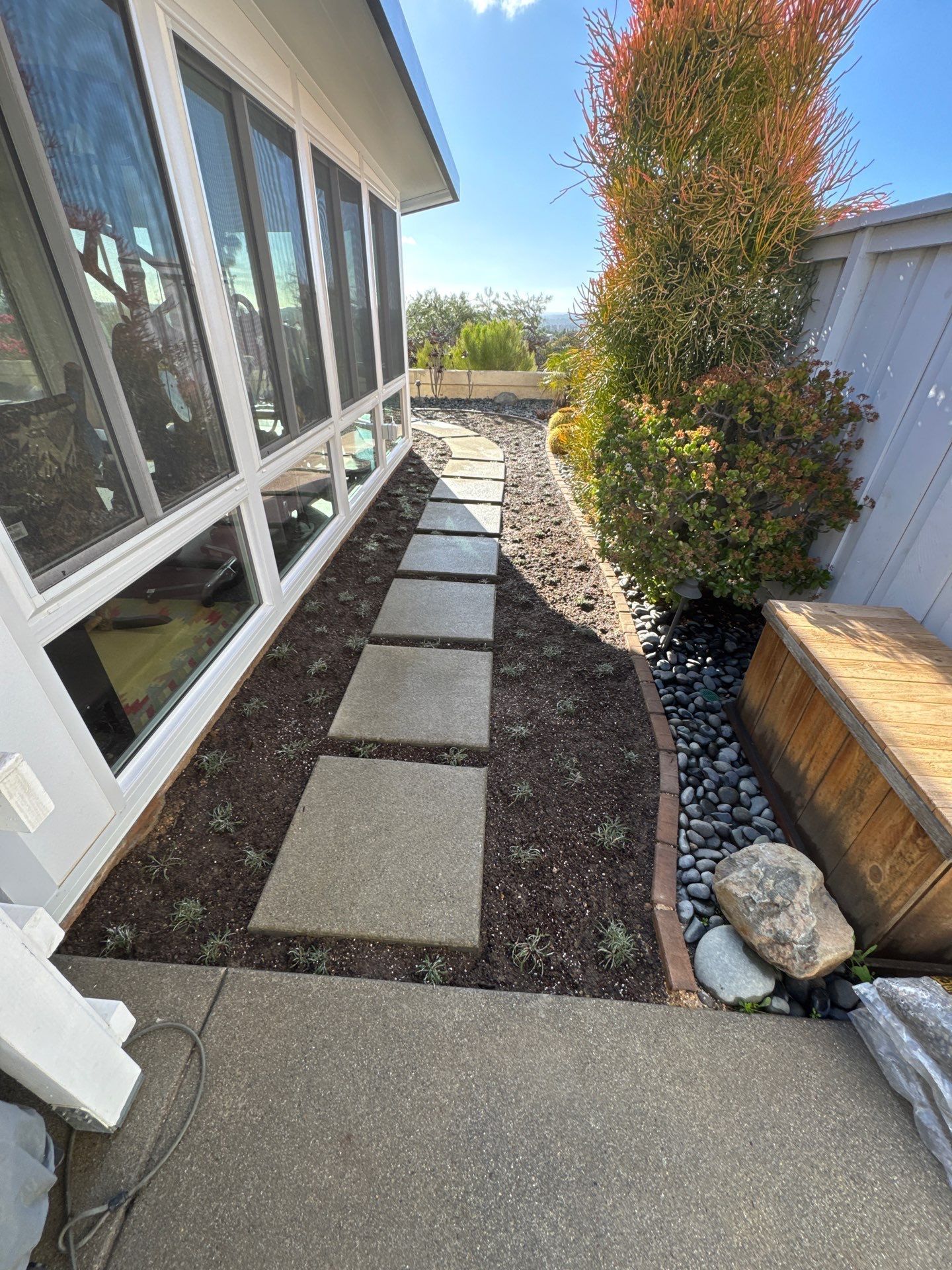 A stone pathway bordered by soil, plants, and a fence, next to a building with large windows.
