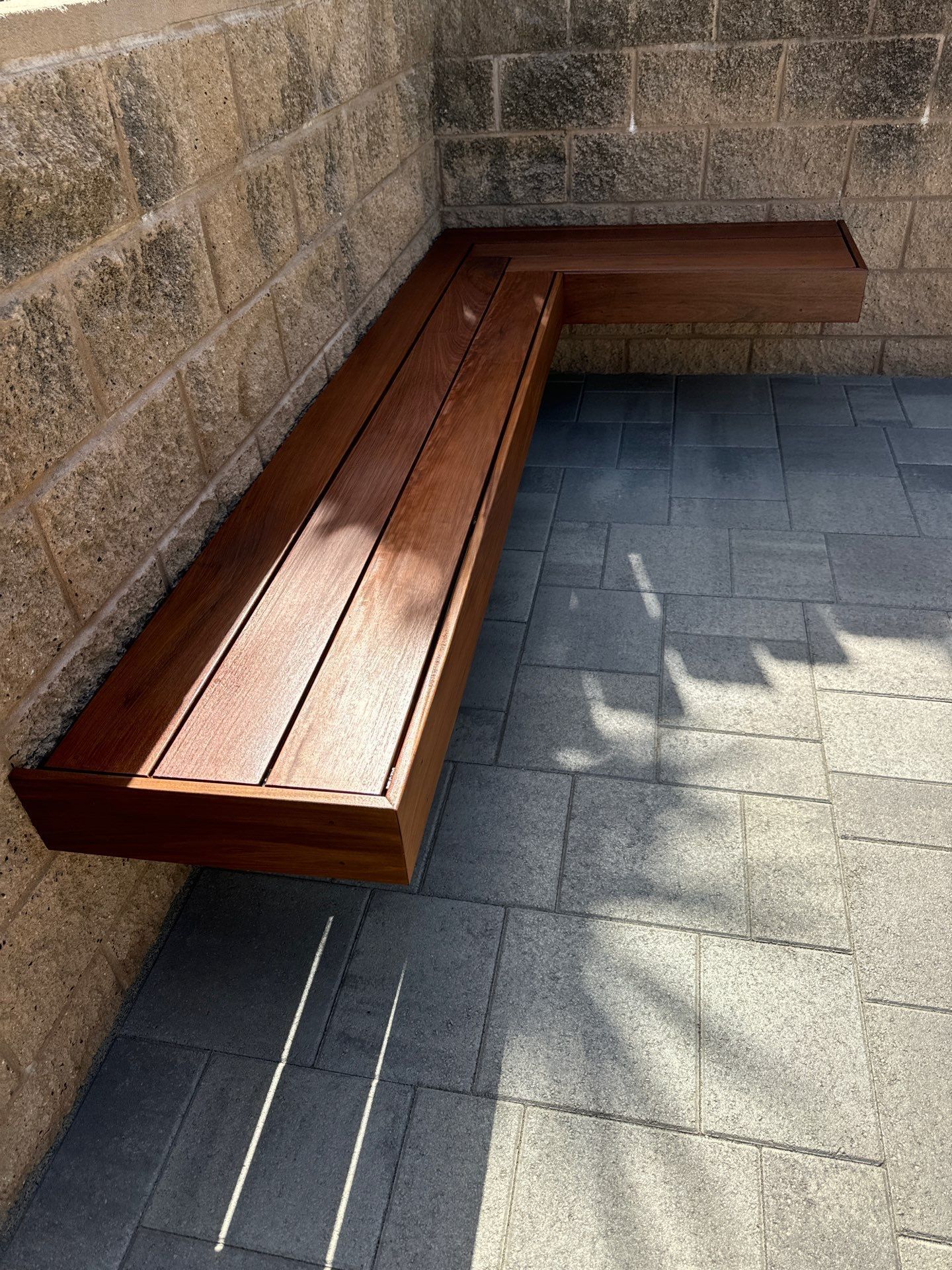 L-shaped wooden bench attached to a brick wall. The bench is brown against a gray stone patio.