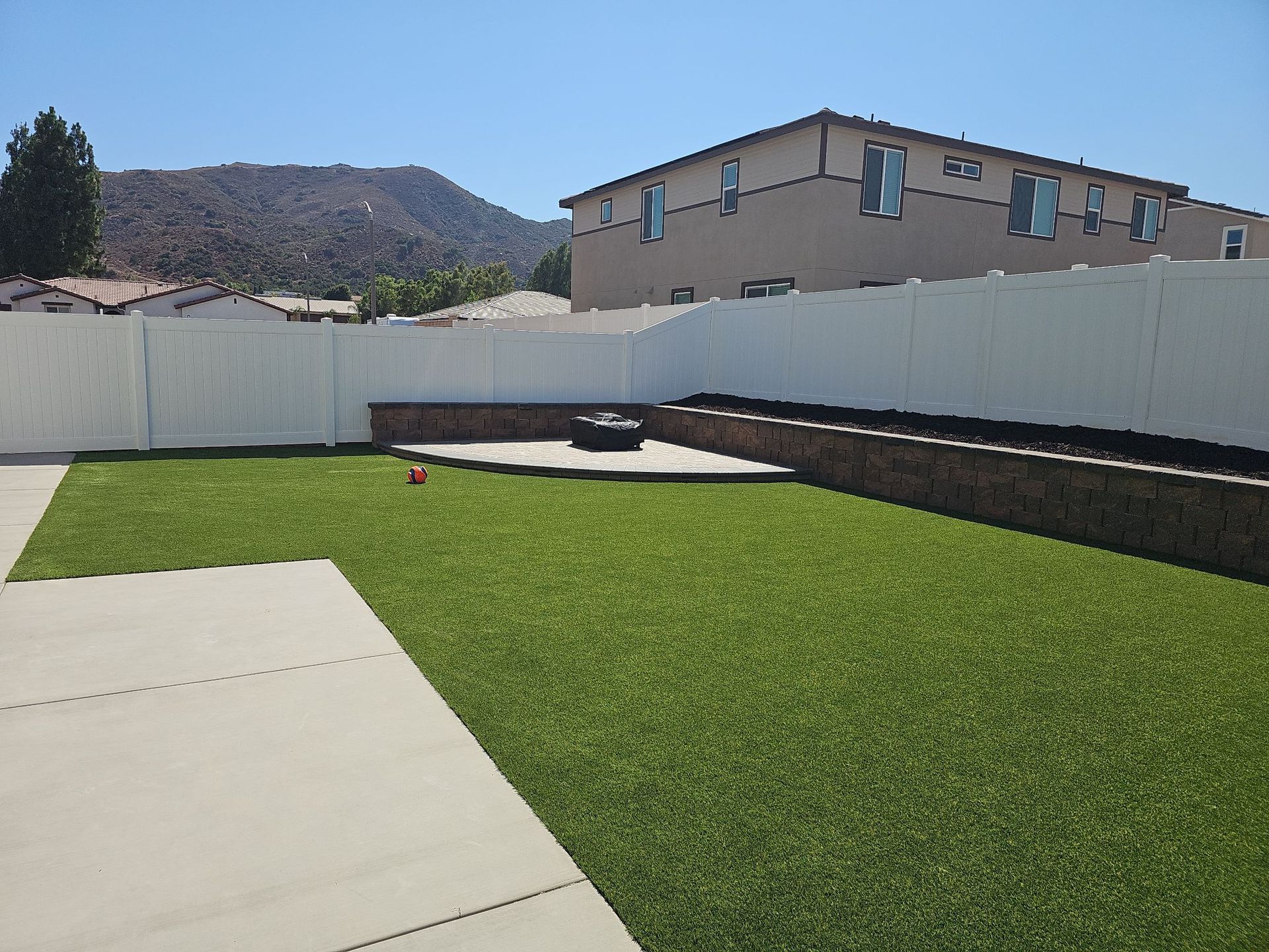 Backyard with green turf, concrete patio, white fence, and a mountain in the background.