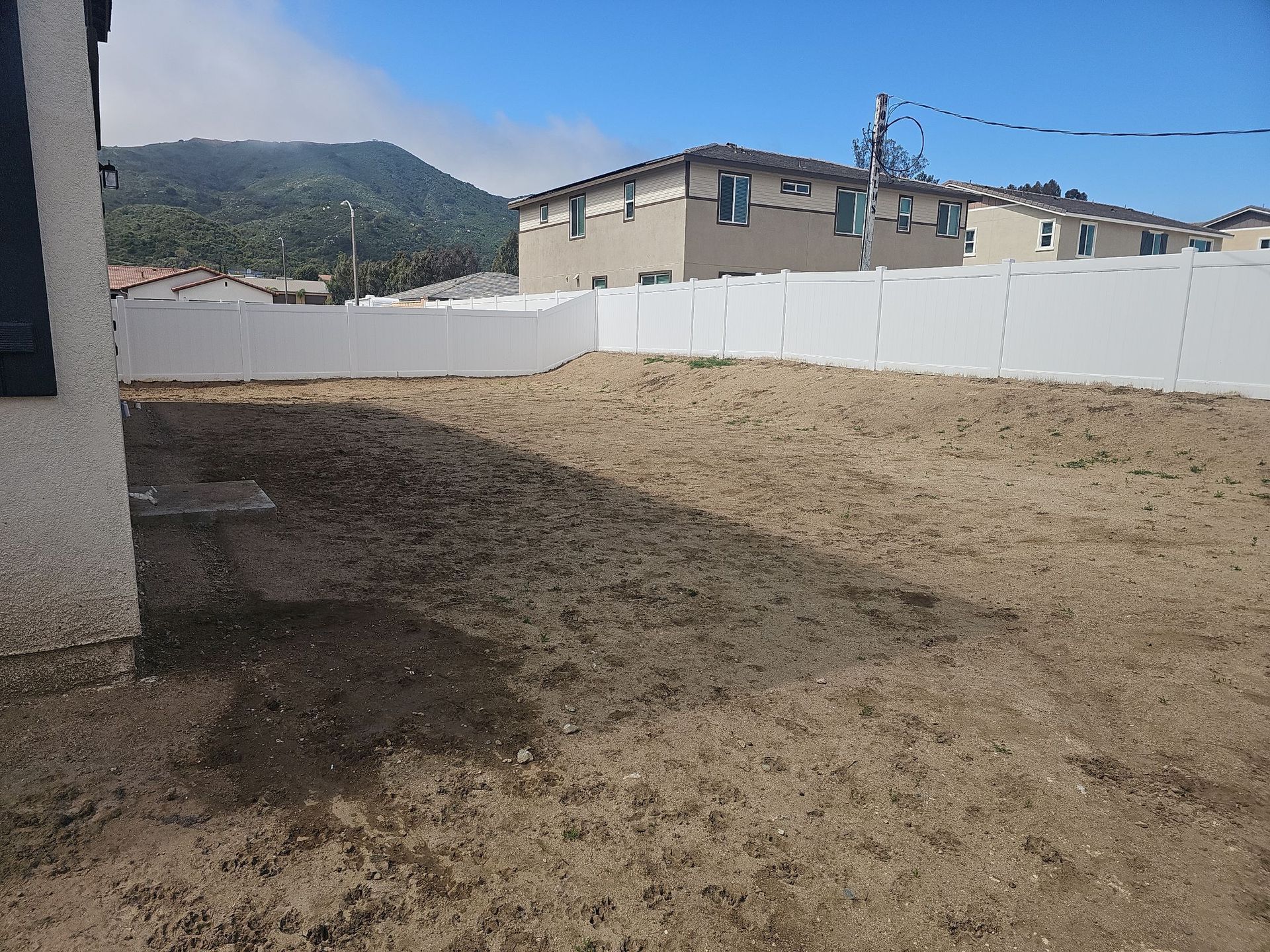 Backyard with dirt ground, white fence, two-story houses, mountain background, and partly cloudy sky.