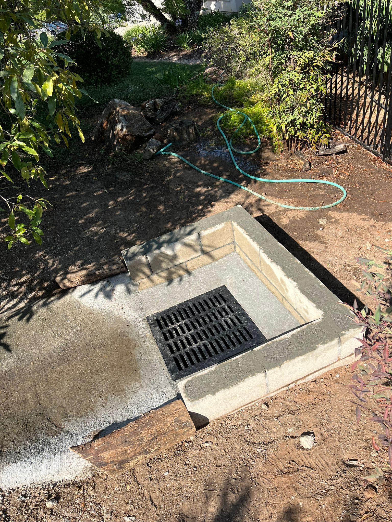 Concrete drainage grate beside a dirt path, with a green hose and fence in the background
