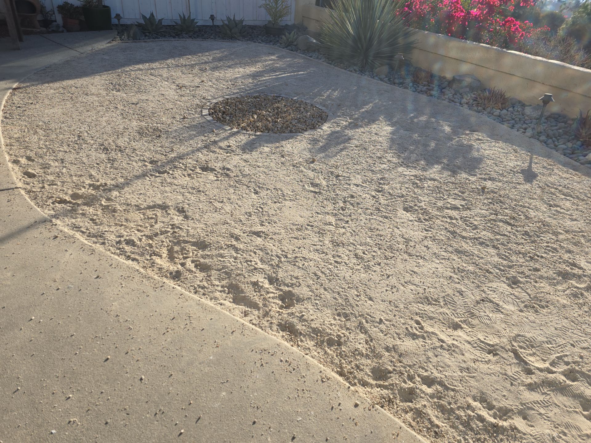 Sandy area next to a concrete walkway, with a circular area of dark rocks and sparse vegetation.