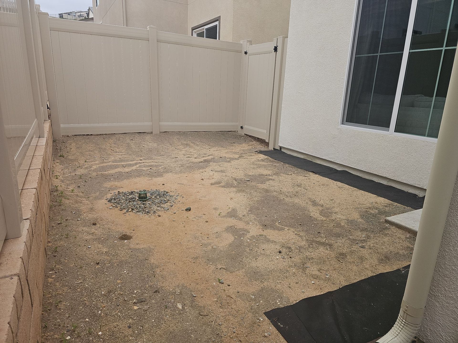 Enclosed backyard with gravel ground, white fence, and a building with a sliding glass door and a downspout.