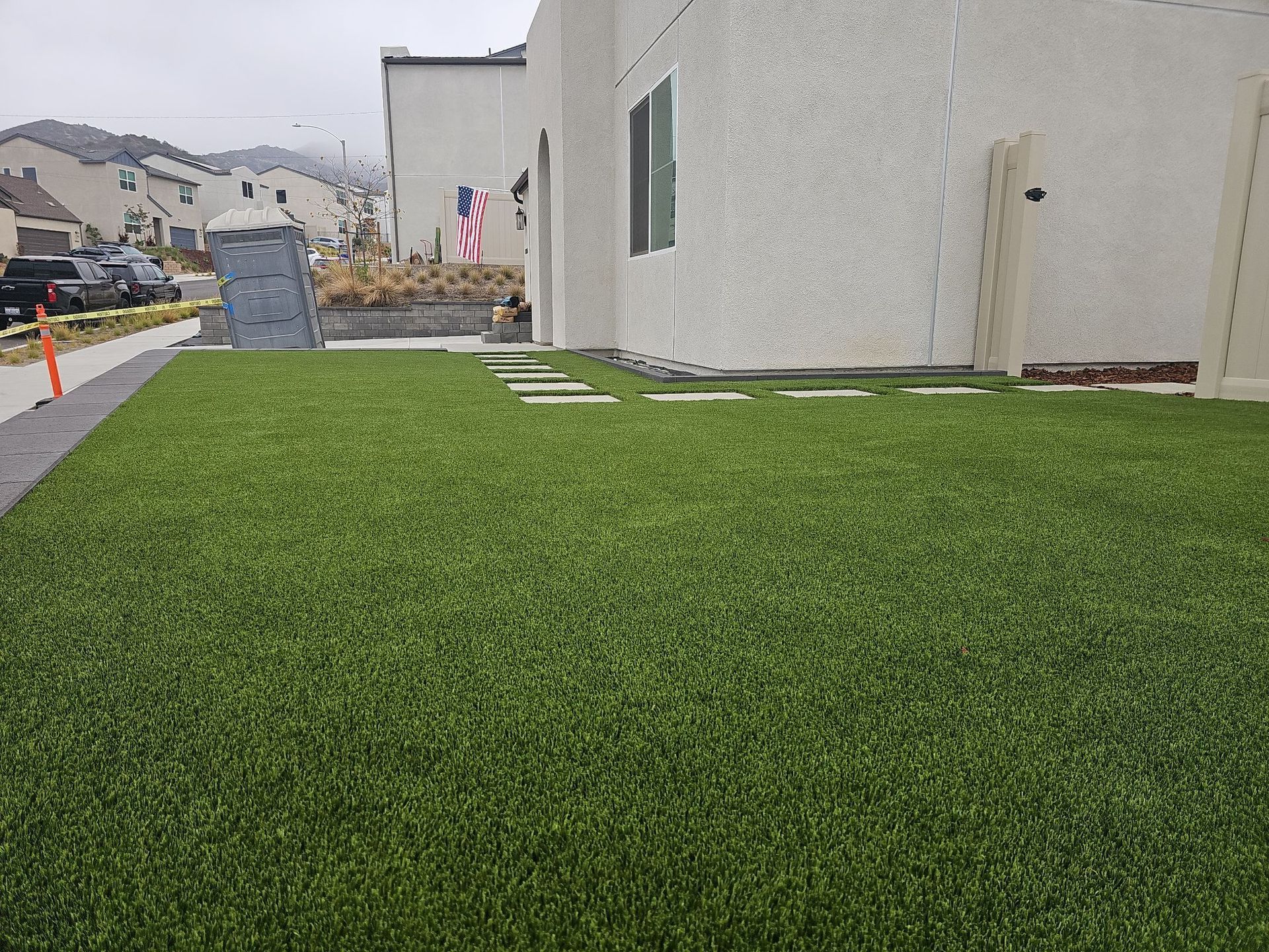 Green artificial lawn in front of a light-colored building with stepping stones and a portable toilet nearby.