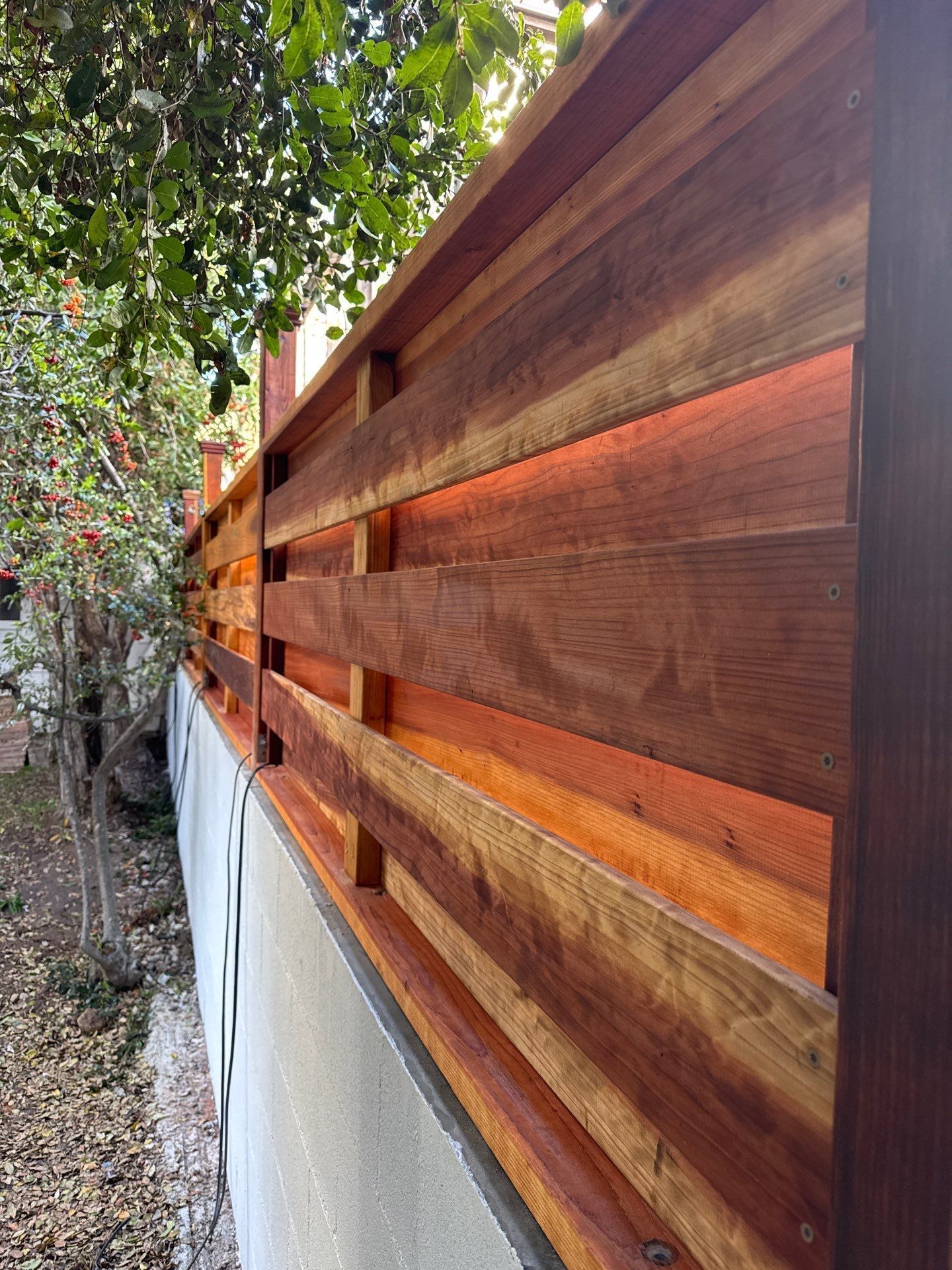 Wooden fence with horizontal planks, dark brown, built on a wall with green foliage above and in the background.