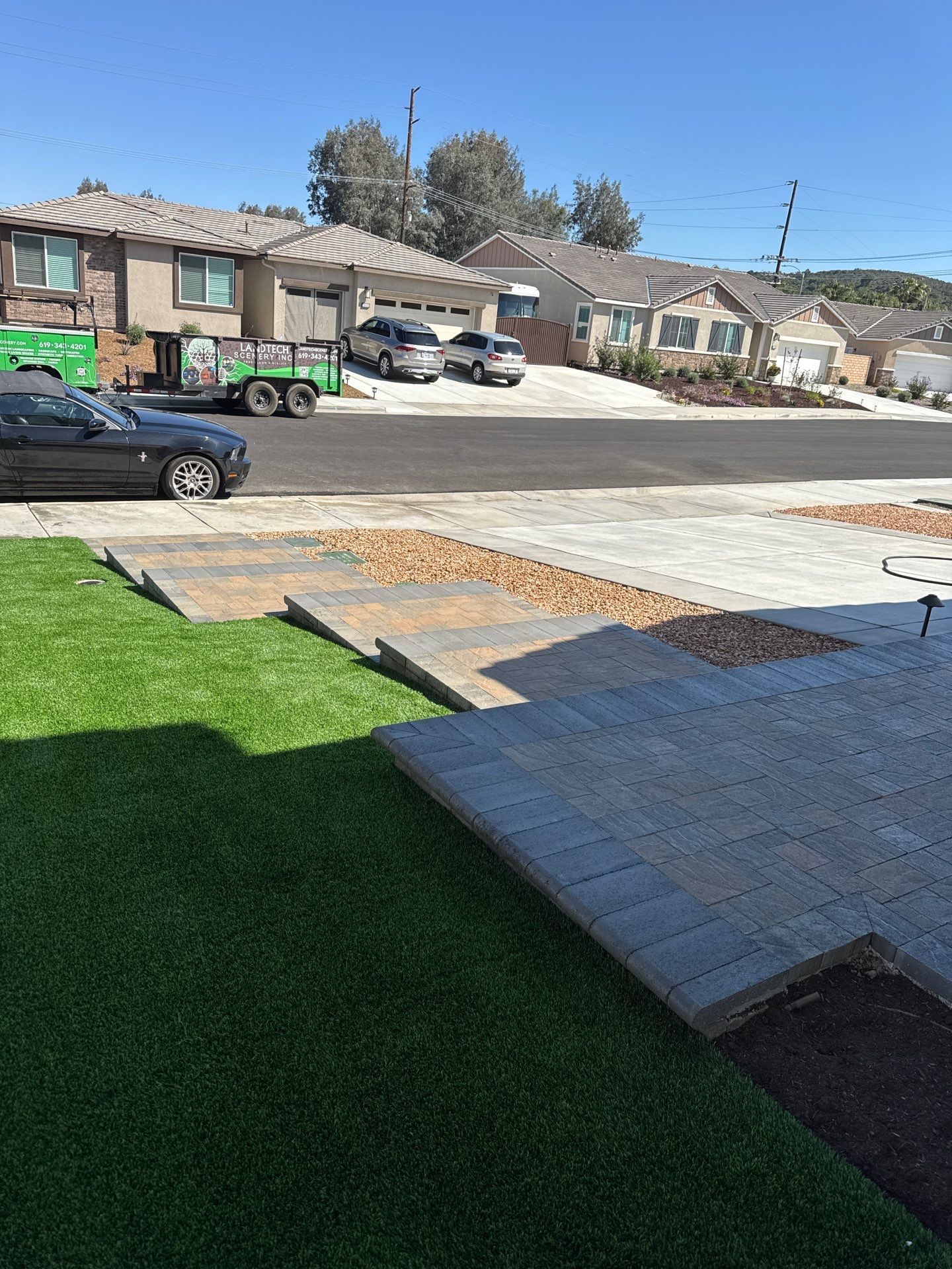 View of a street with newly paved walkways, green lawns, and houses under a clear blue sky.