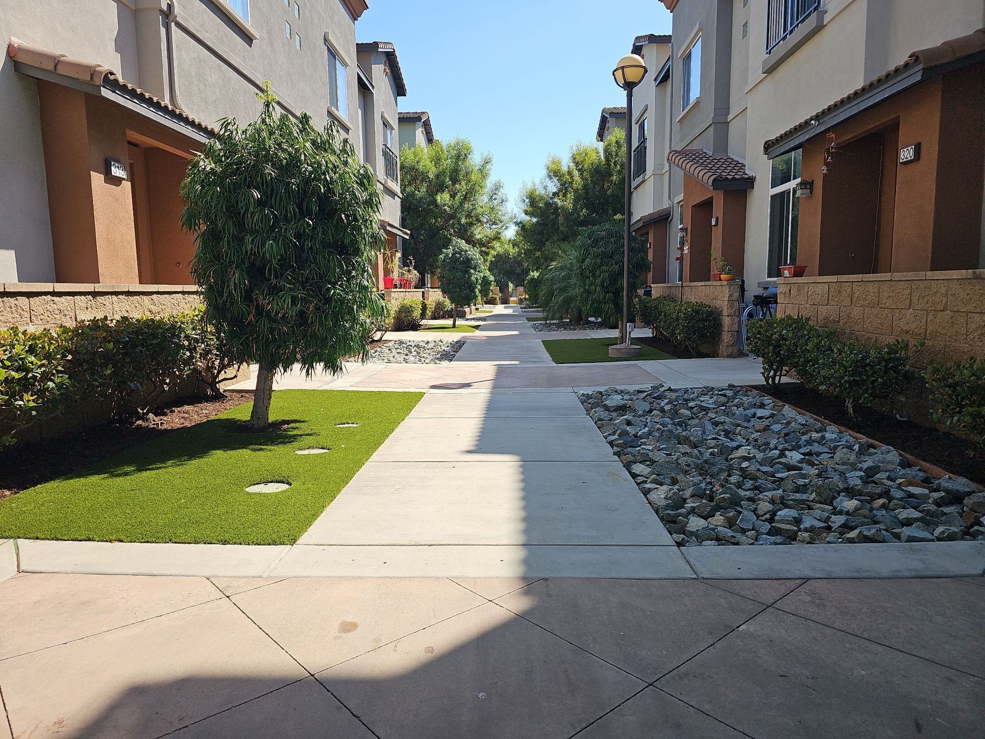 Sidewalk in a residential area. Beige buildings Project path. Landscaping includes grass, trees, and rocks.