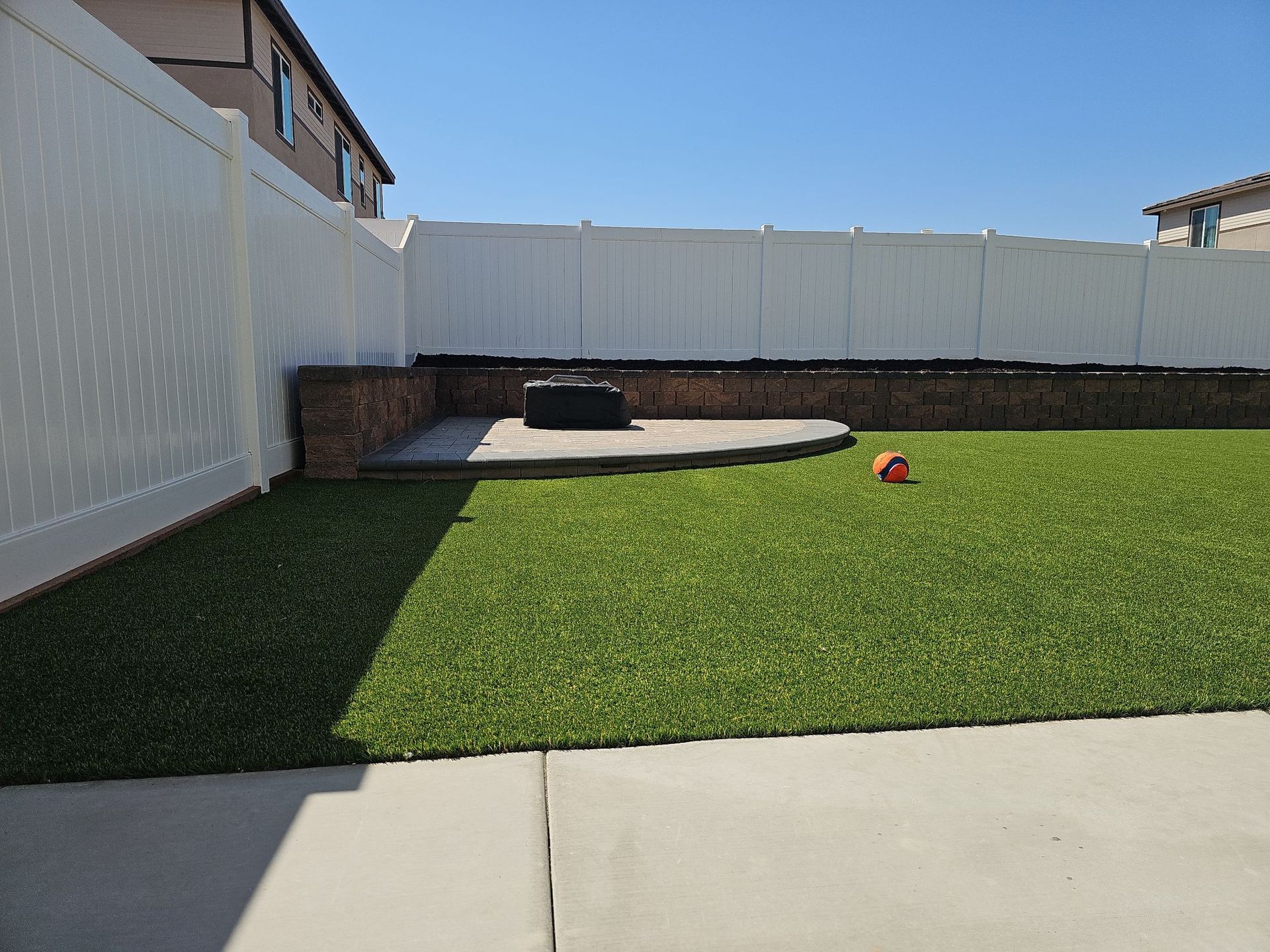 A backyard with green turf, a white fence, and a stone fire pit. A ball sits on the grass.