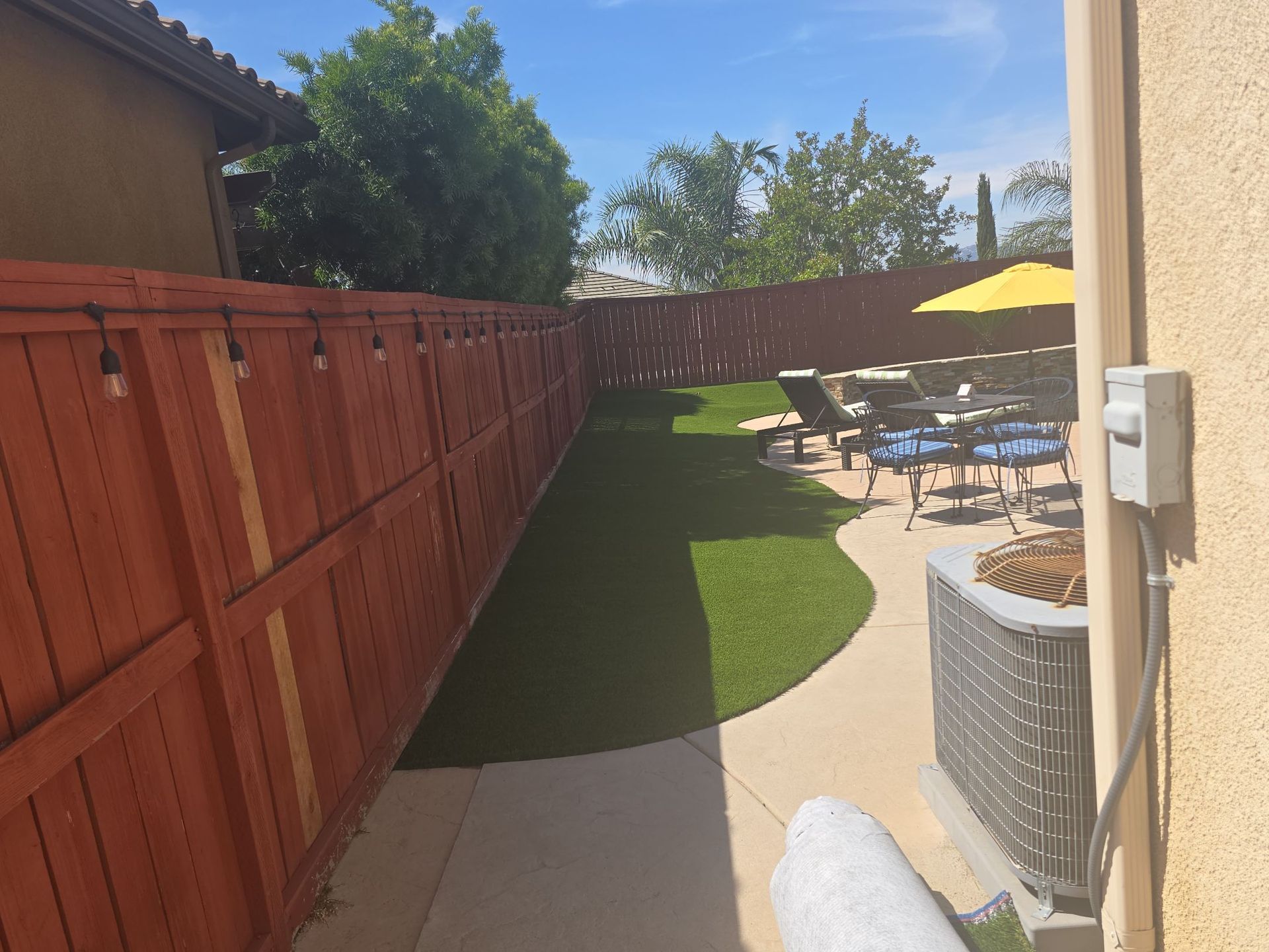 Lush green lawn with red wooden fence, pathway, outdoor seating area with yellow umbrella, blue sky.