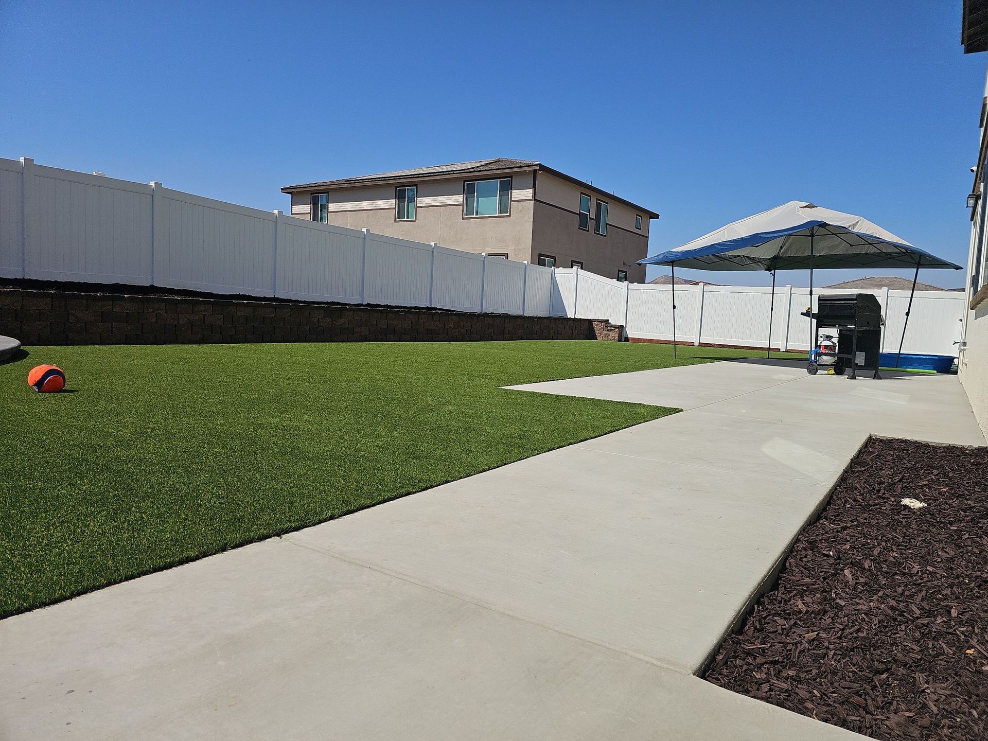 Backyard with artificial grass, concrete patio, white fence, barbecue grill, and house.