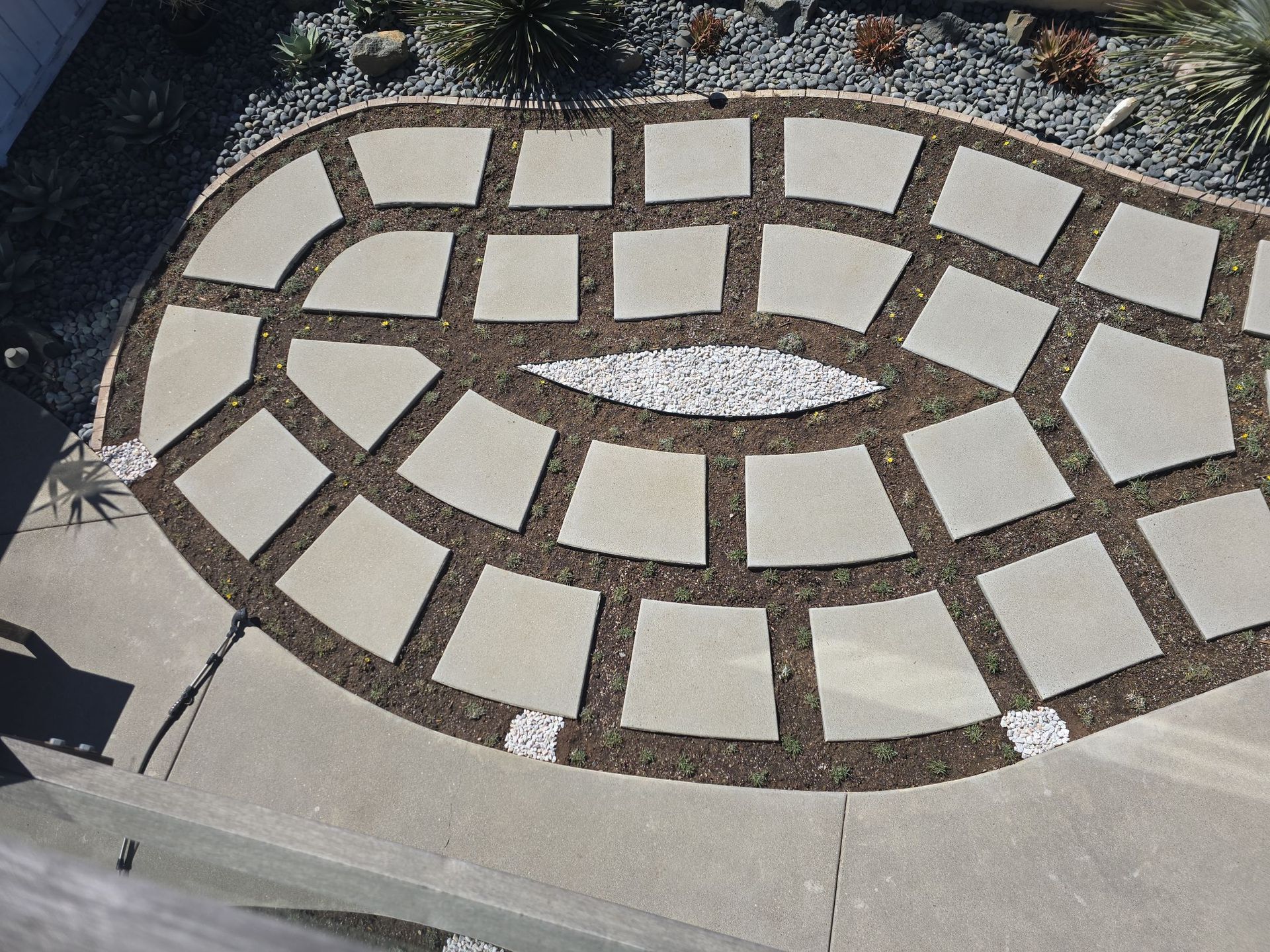 Stone paver patio with a central oval of pebbles, surrounded by mulch and bordering a concrete surface.