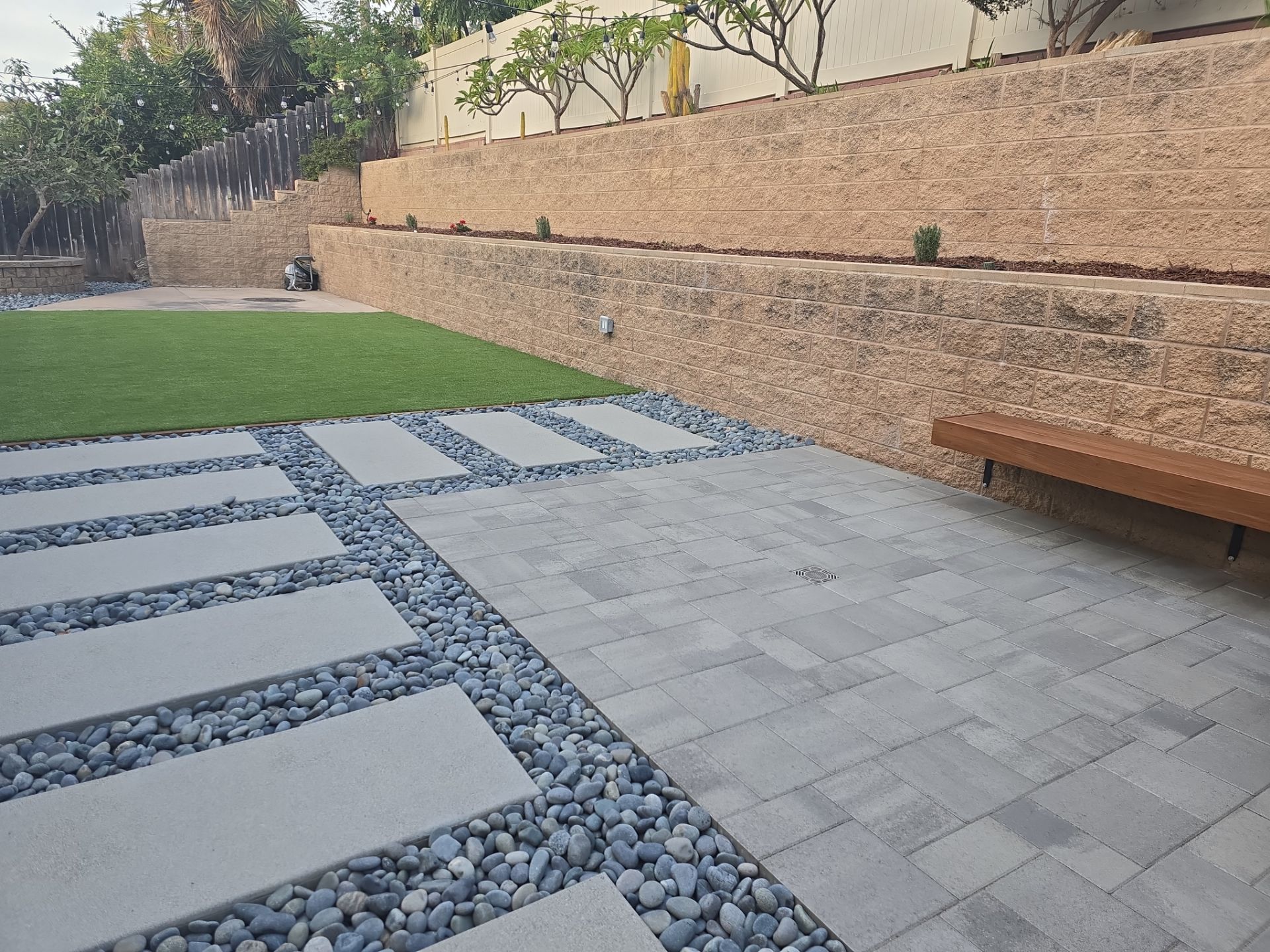 Patio with stone pavers, dark pebbles, and a retaining wall in a backyard setting.